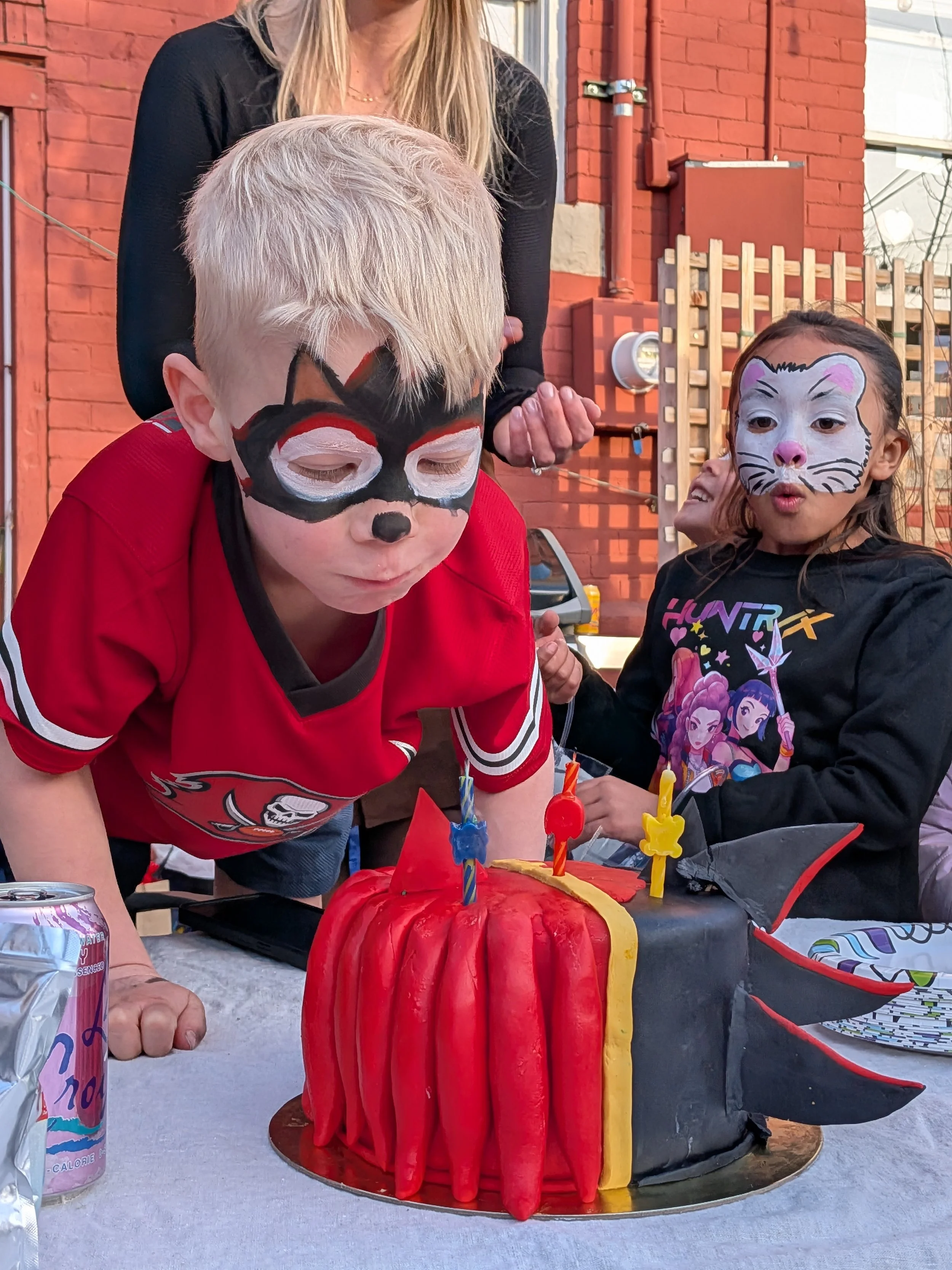 Children with face paint at a birthday party, one dressed as a cat and the other as a mouse, blowing out candles on a red and black Halloween-themed cake decorated with Bat and spider motifs, outdoors in a backyard.
