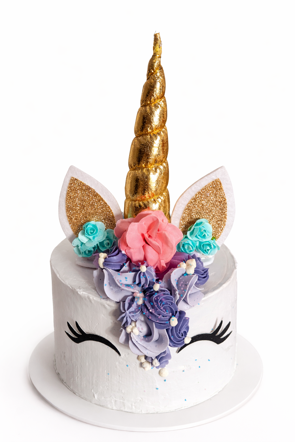 A unicorn-themed cake with a gold unicorn horn, glittery ears, black eyelash decorations, and pastel-colored flowers.