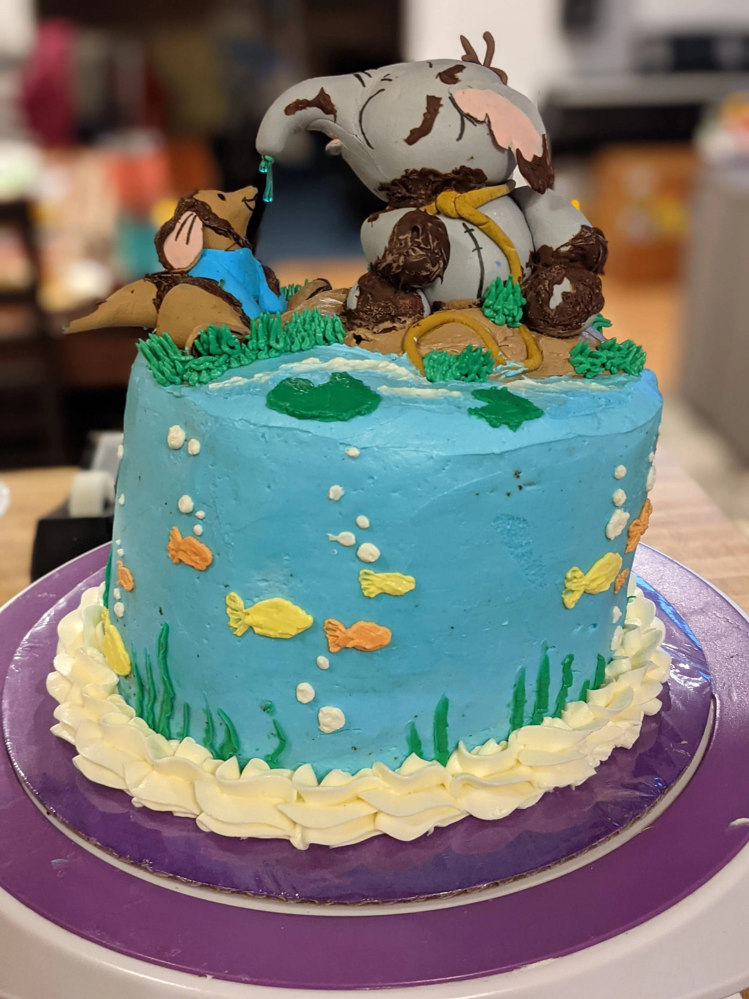 A birthday cake with an underwater ocean theme, decorated with blue frosting, fish, seaweed, and an edible figurine of a bear and a mouse. The figurines are depicted in a playful scene on top of the cake.