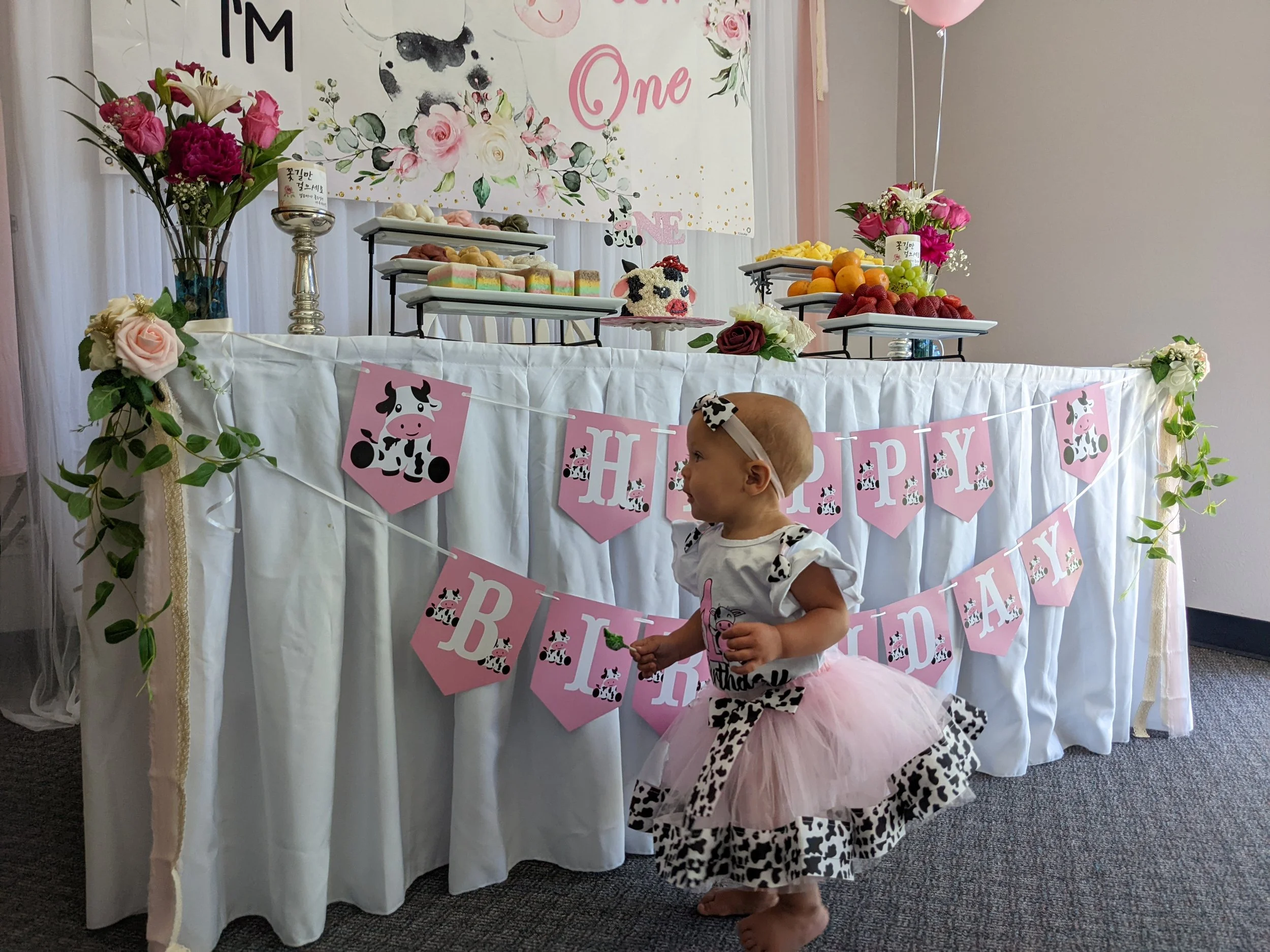 A young girl in a cow-themed dress and pink tulle tutu stands in front of a decorated birthday table with pink and cow-themed accents, including a "Happy Birthday" banner, floral arrangements, and tiered trays of fruits and sweets, at a birthday cele