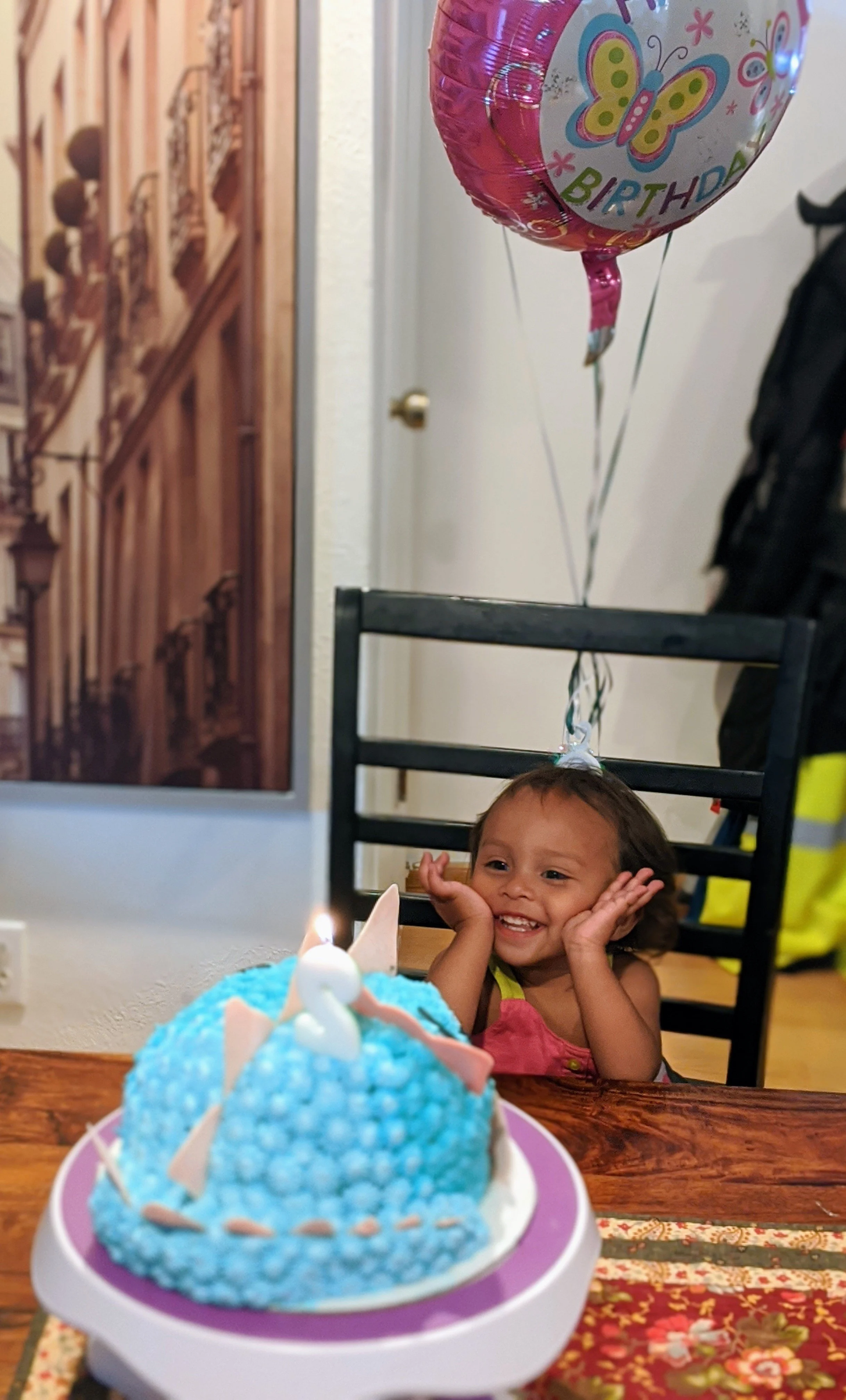 A young girl sitting at a birthday celebration, smiling and resting her hands on her cheeks, with a colorful birthday cake with a lit candle in front of her. A pink butterfly-themed birthday balloon floats above her.