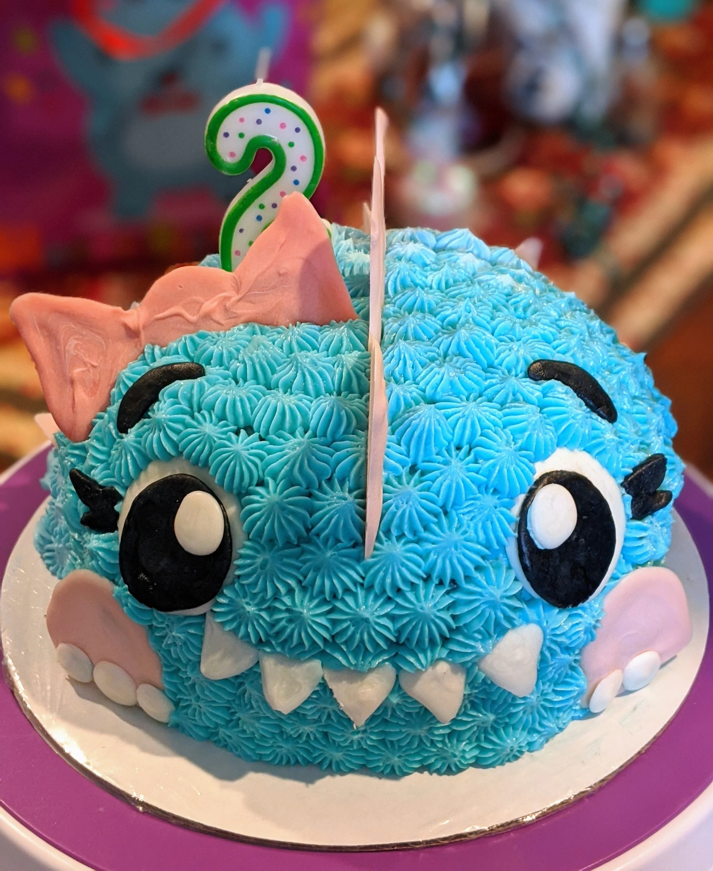 A colorful birthday cake decorated to look like a blue, fluffy, cartoon monster with large black and white eyes, pink ears, and small white teeth. A green and white number 2 candle is on top.