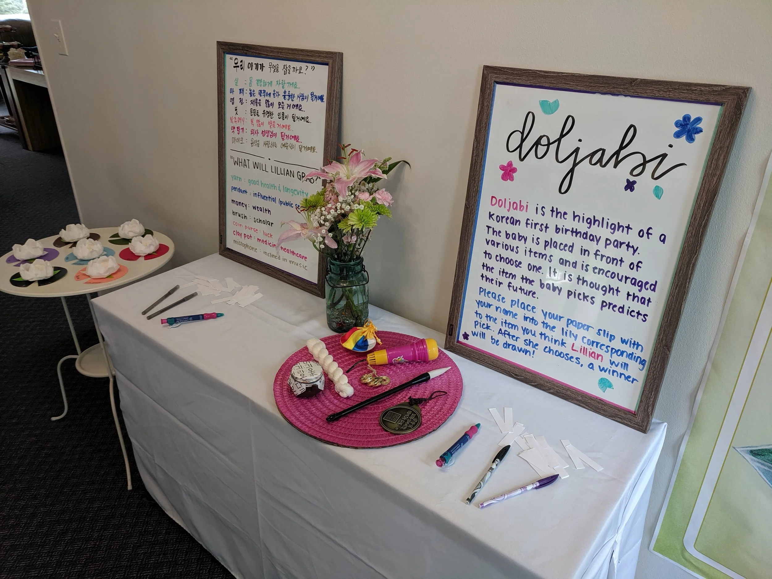 Table with white tablecloth displaying decorations for a baby shower or birthday party with two framed posters, flowers in a vase, pens, and a pink woven placemat with items including a lollipop, whistle, and medal.