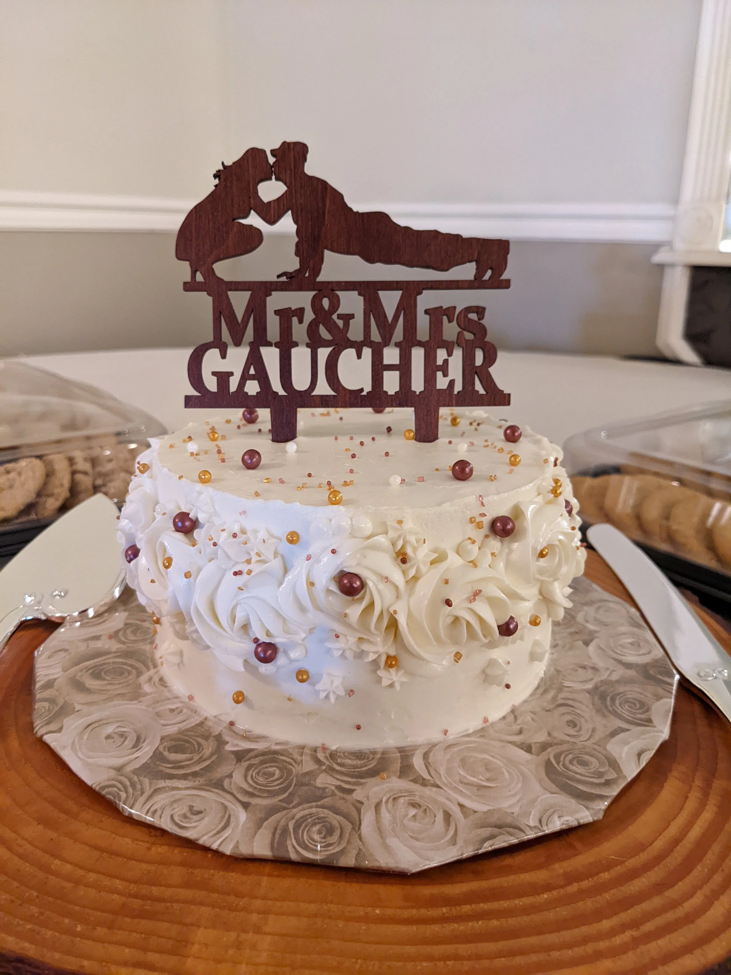 A round white wedding cake with piped rosettes and small decorative beads, topped with a wooden silhouette cake topper that says 'Mr & Mrs Gaucher' and features a couple in a romantic pose.