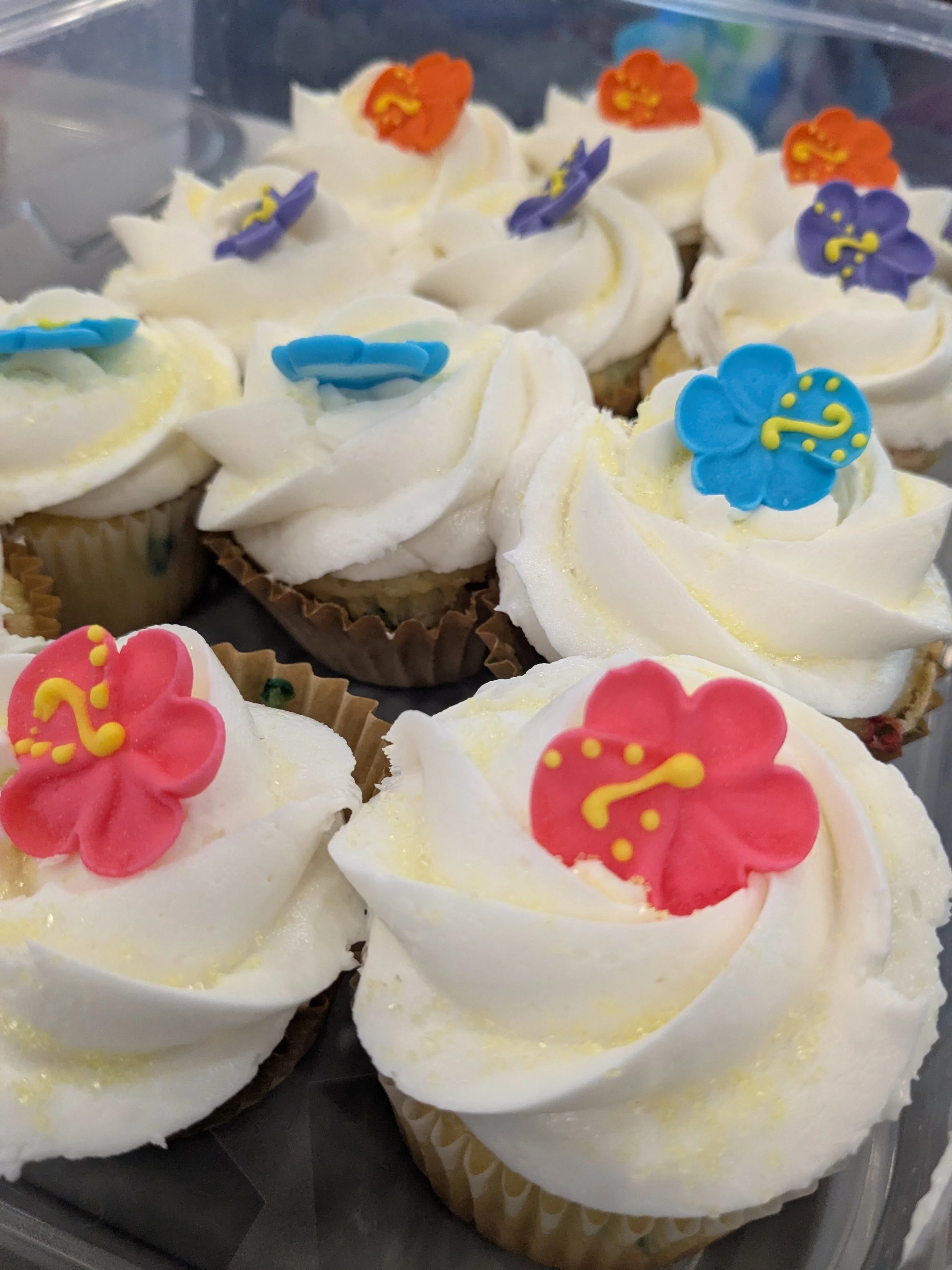 Cupcakes with white frosting decorated with colorful flower-shaped edible toppers in pink, blue, purple, and orange.