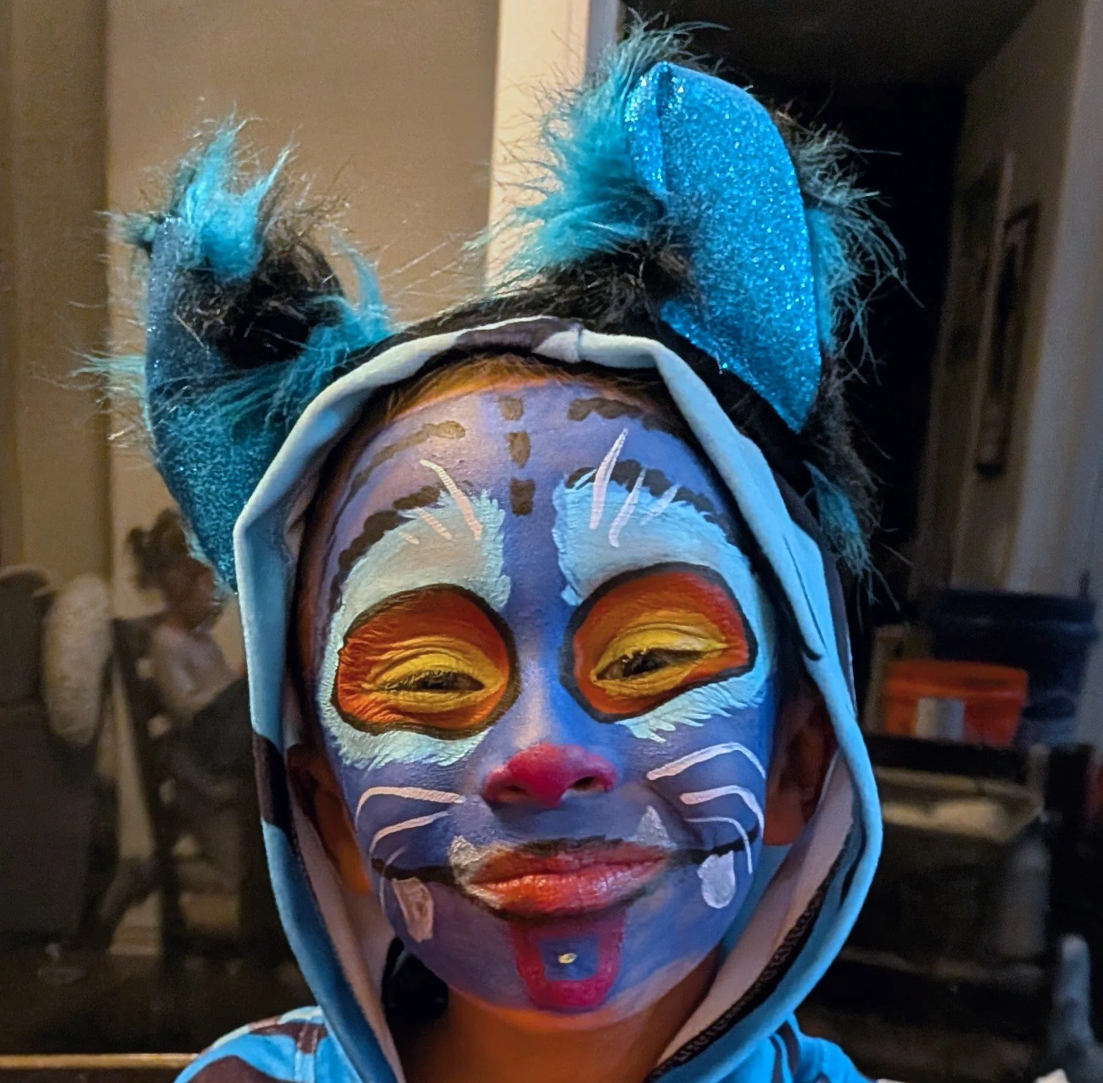 Child with face paint resembling a cat, wearing a blue dinosaur hat with ears, indoors with other children in the background.