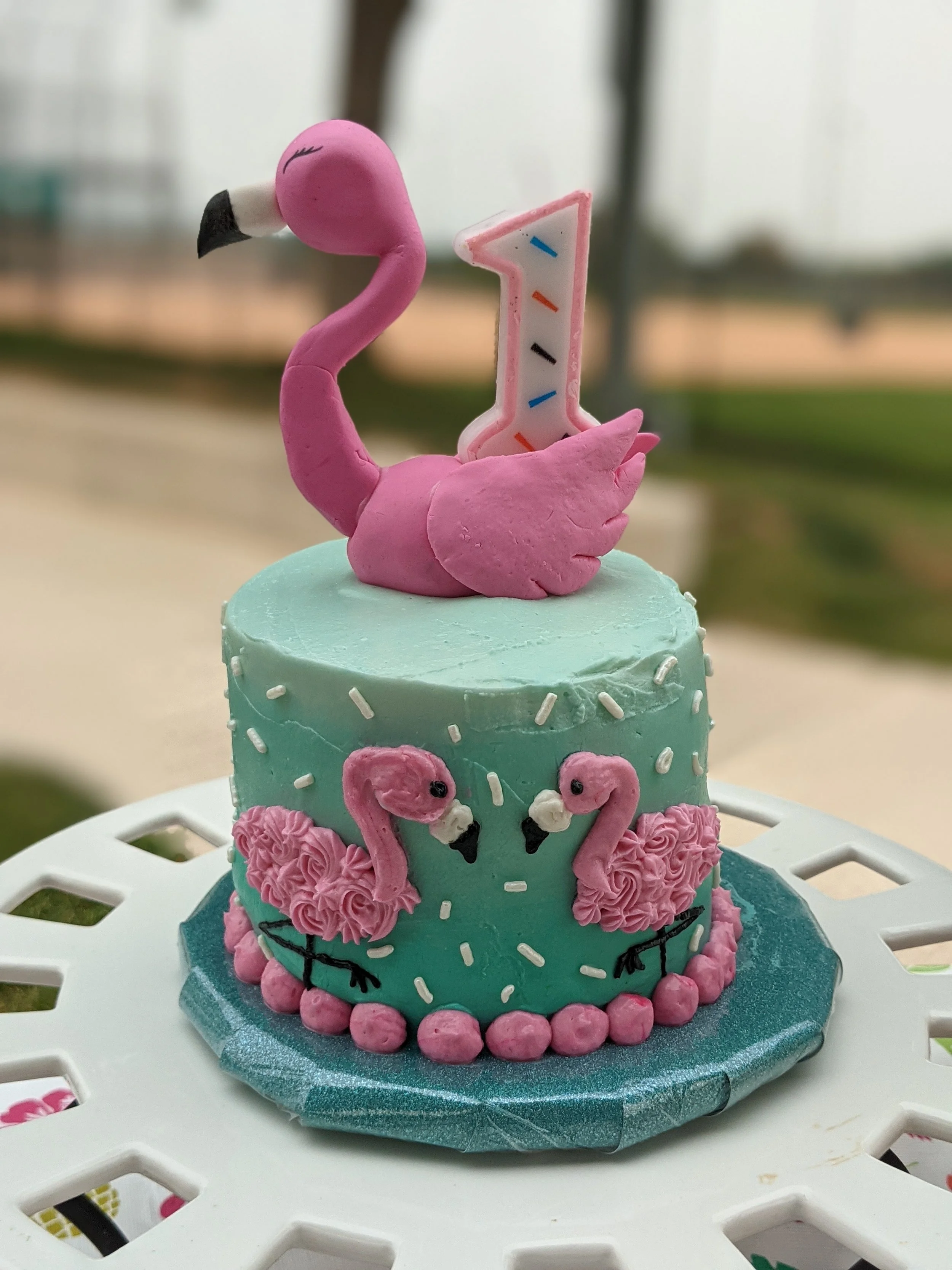 A birthday cake decorated with pink flamingos, a large number one candle, and pastel colors, set on a white table outside.