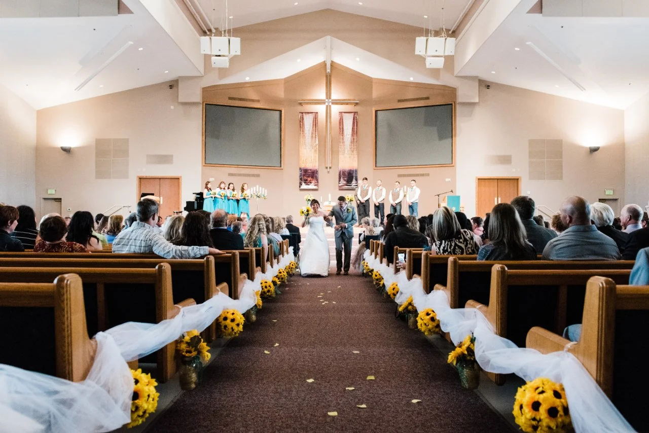 A wedding ceremony inside a church, with a bride and groom walking down the aisle, surrounded by guests, floral decorations, and a cross at the altar.