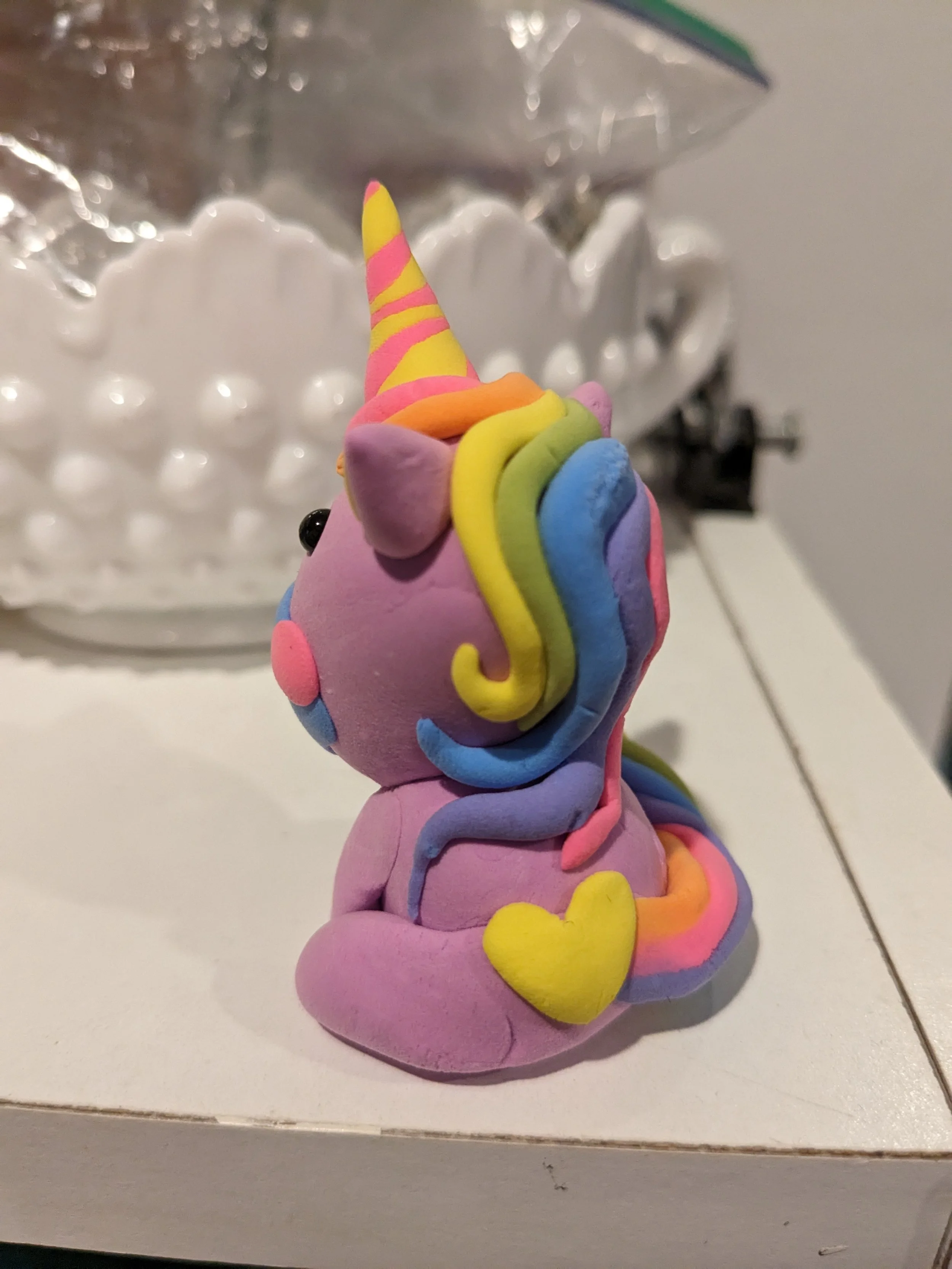 Colorful figurine of a cartoonish unicorn with a rainbow mane and tail, sitting on its hind legs with a yellow heart on its side.