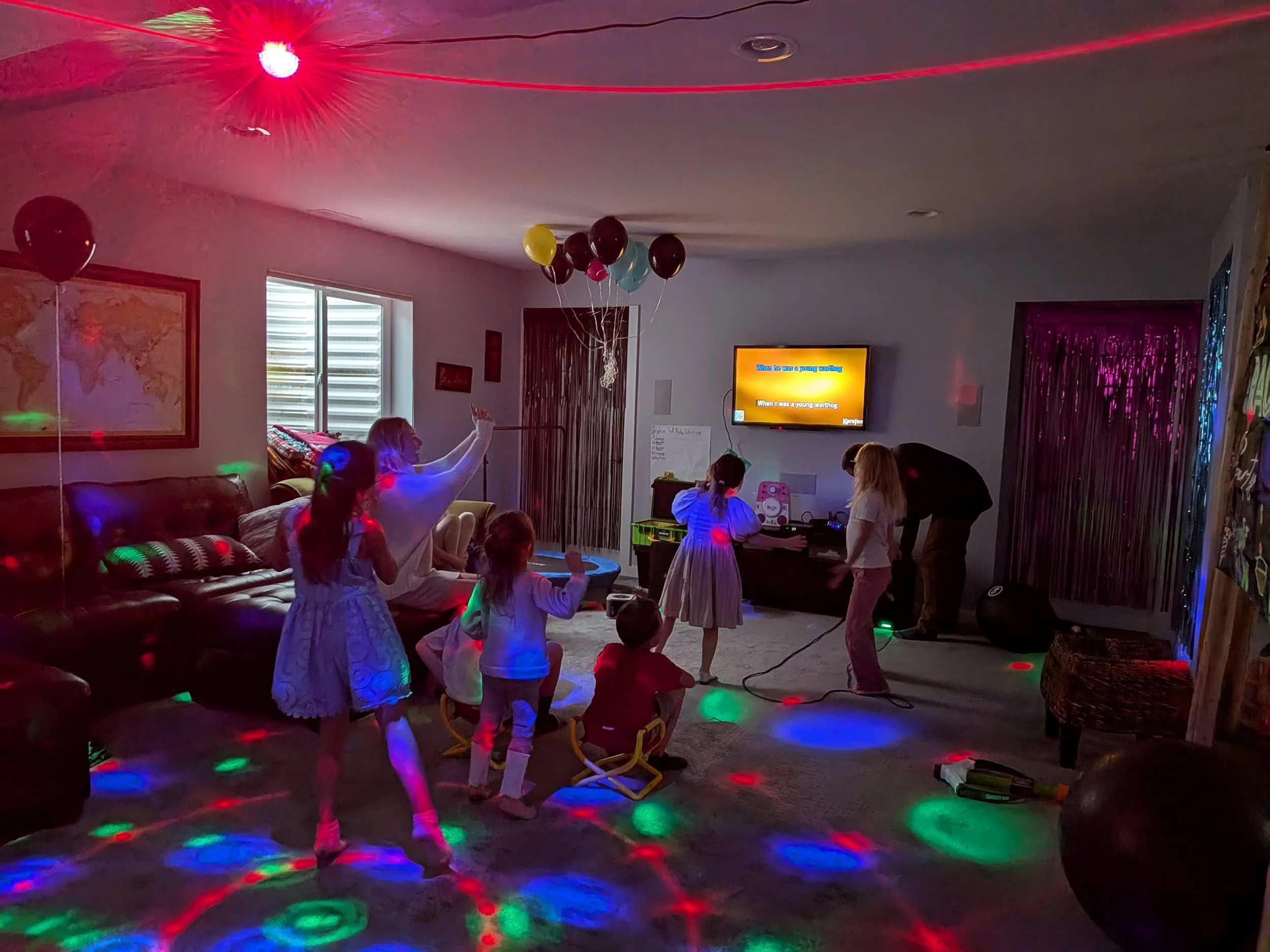 Kids and adults dancing and playing with balloons in a living room with colorful disco lights, a television screen, and a map on the wall.