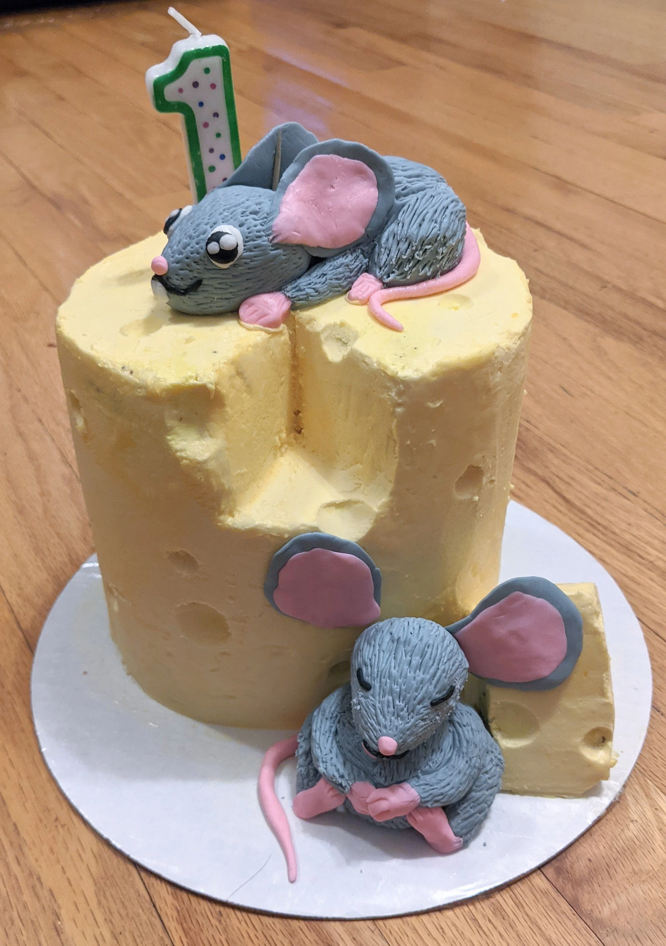 A birthday cake decorated with two gray mice figurines, one on top and one at the base, with pink ears and tails. The cake has a yellow frosting with holes, and a candle shaped like the number one on top.