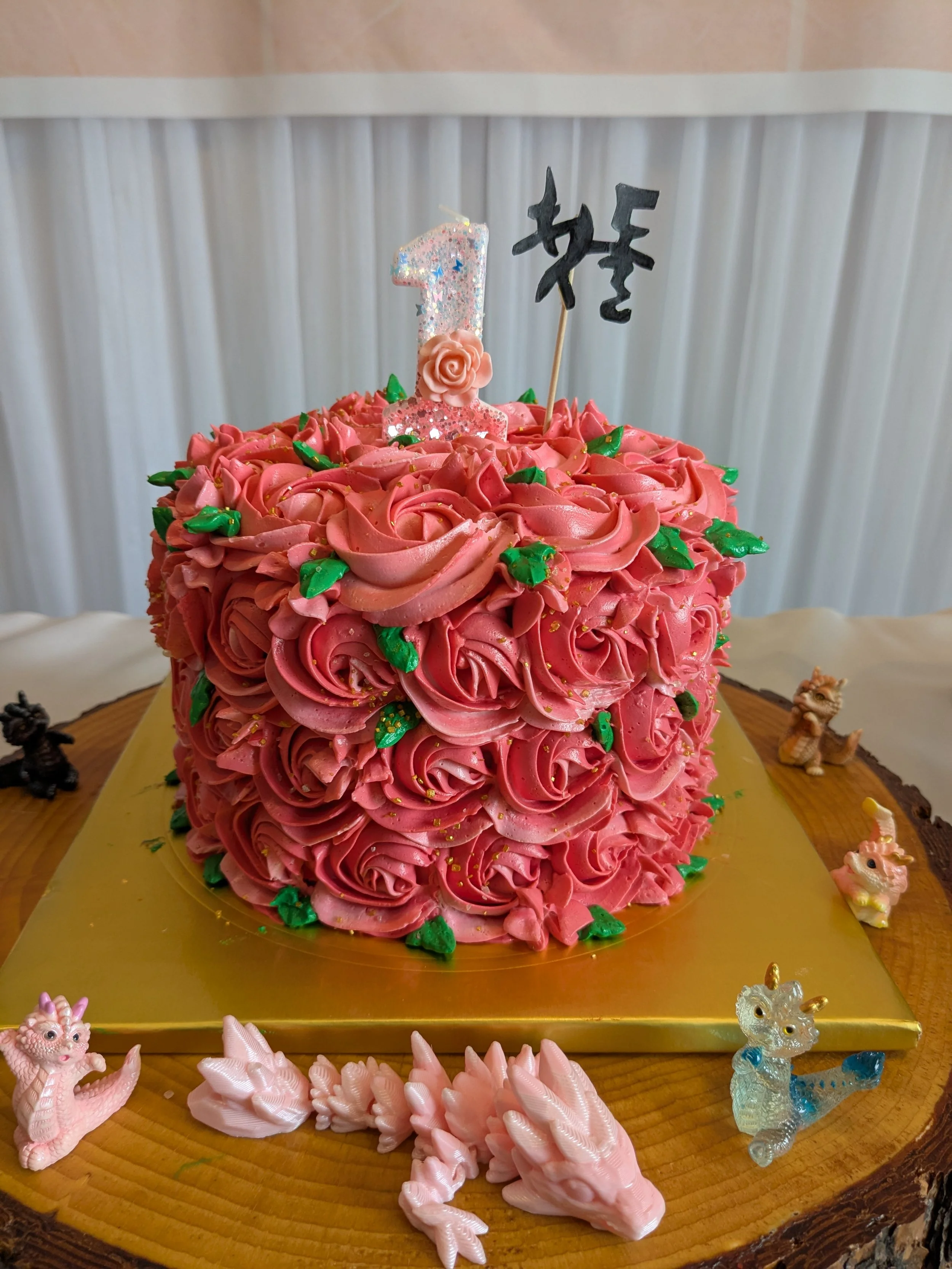 Pink roses decorated birthday cake with a glittery number one candle, a black Seoul sign topper, surrounded by small dragon and unicorn figurines on a wooden table.