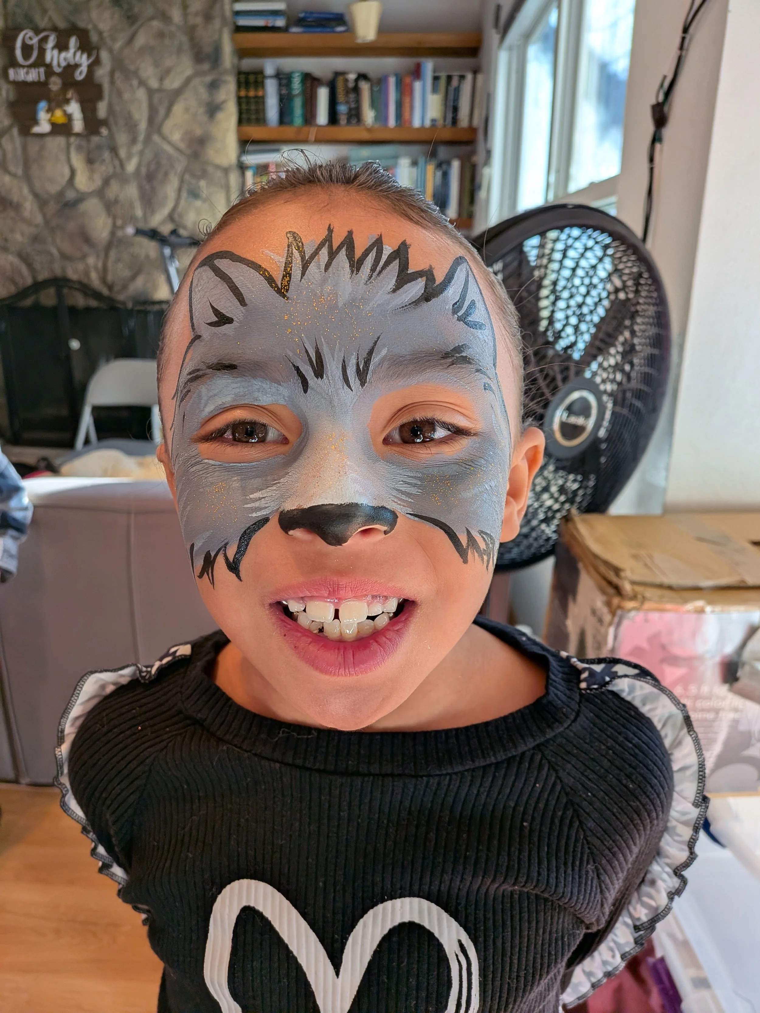 Child with a face painted as a wolf, smiling indoors with bookshelves and a fan in the background.