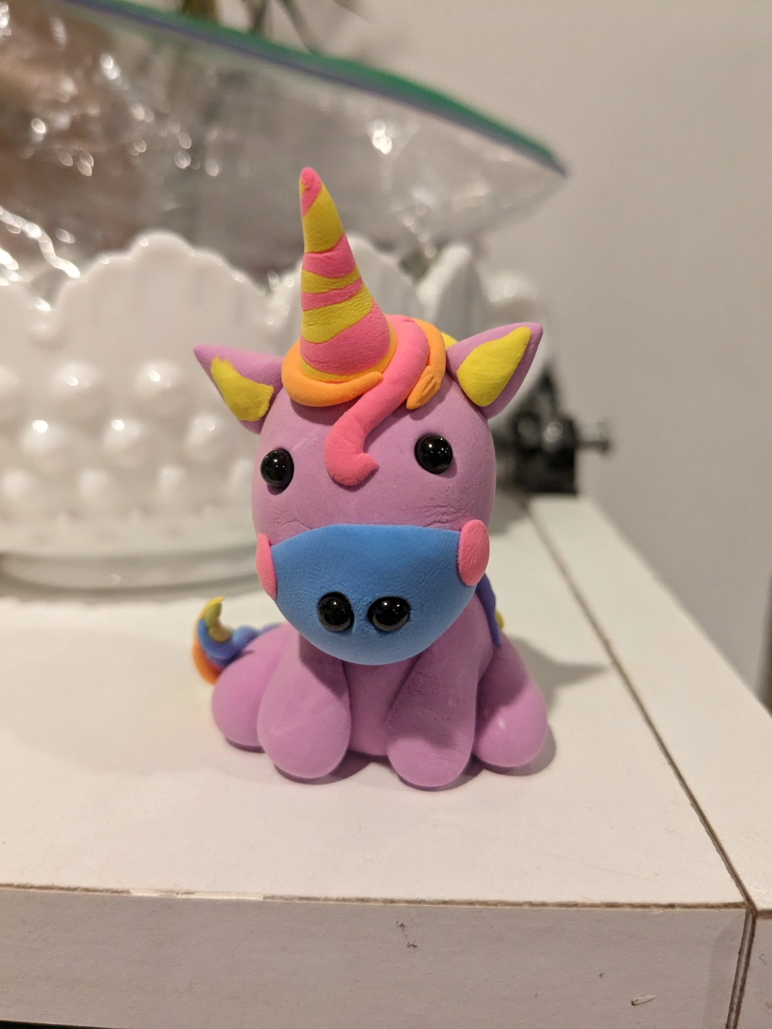 A small, colorful unicorn figurine made of clay or similar material, with a pink body, a striped yellow and pink horn, a multicolored tail, and a blue face mask, sitting on a white tiled surface.