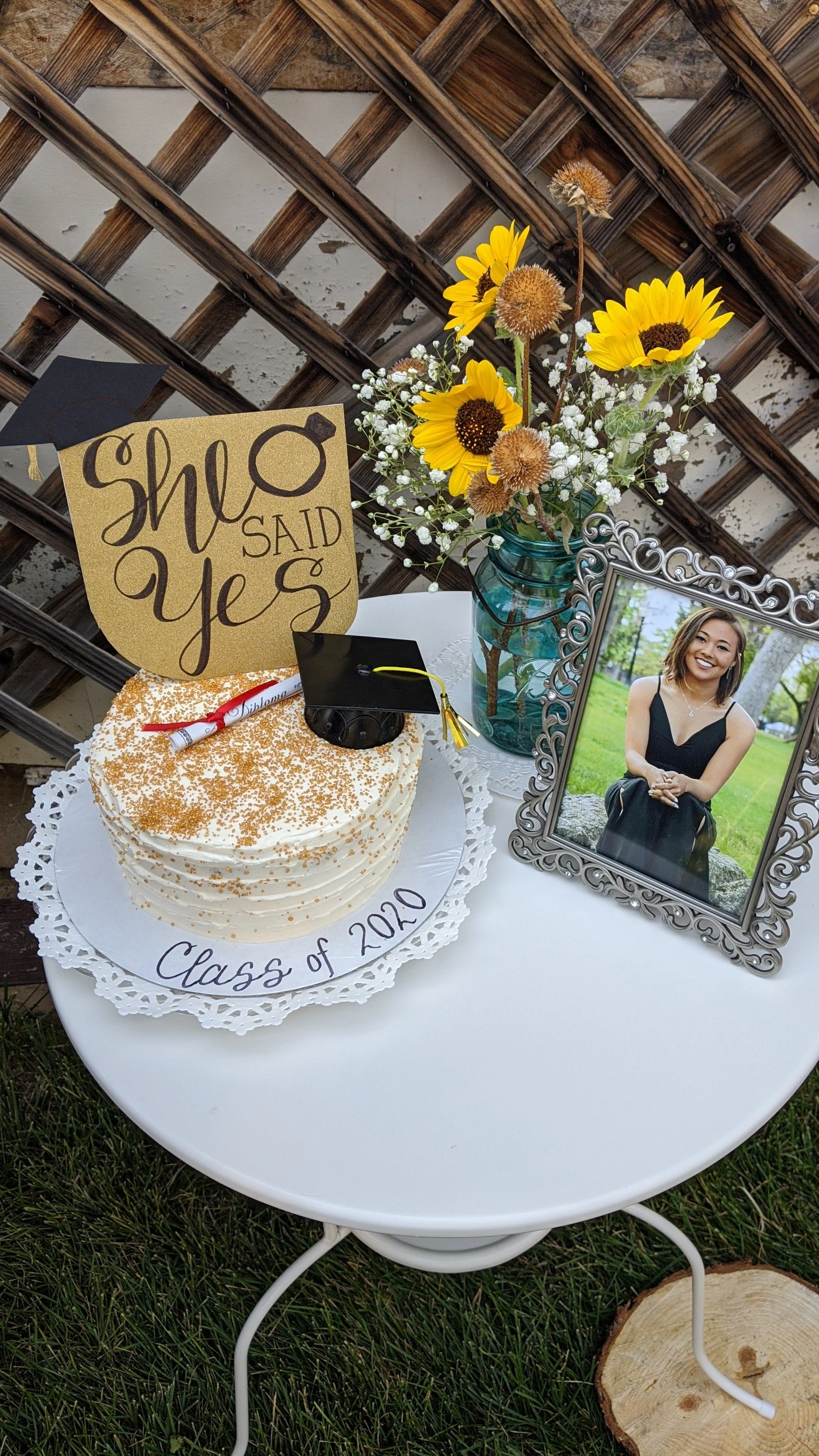 A graduation celebration setup with a decorated cake, a framed photo of a woman, a bouquet of sunflowers in a glass jar, and a sign that says 'She Said Yes.' The cake has a gold star and a graduation cap ornament, and the table has a paper doily and 