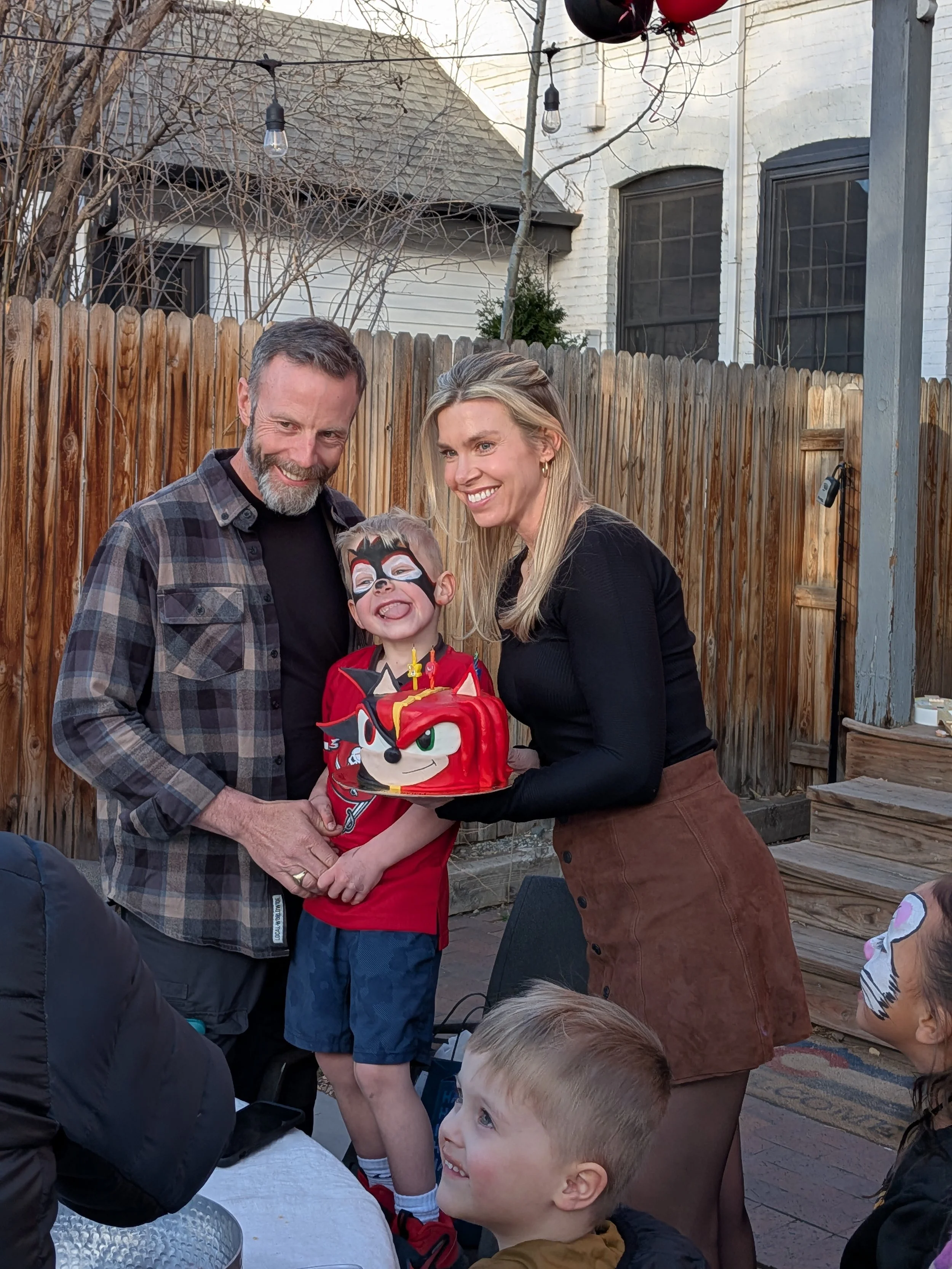 A young boy with face paint and a red Sonic the Hedgehog themed birthday cake is smiling while standing between two adults, an older man and a woman, who are holding the cake together. The scene is outdoors in a backyard with a wooden fence and strin