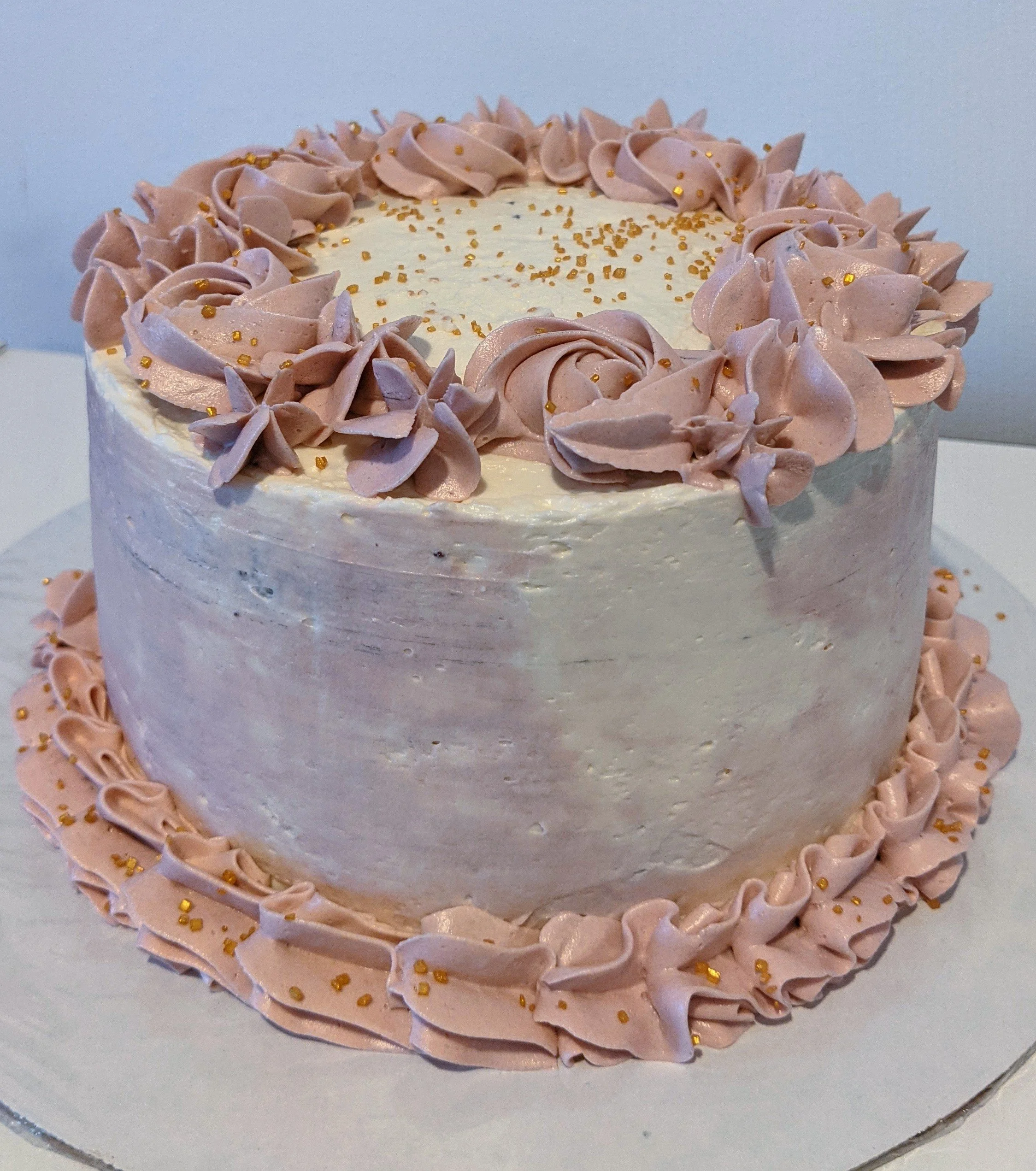 A round cake decorated with pink frosting and gold sprinkles. The cake has a pink frosting border on the top and bottom edges and pink rosette decorations on the top.