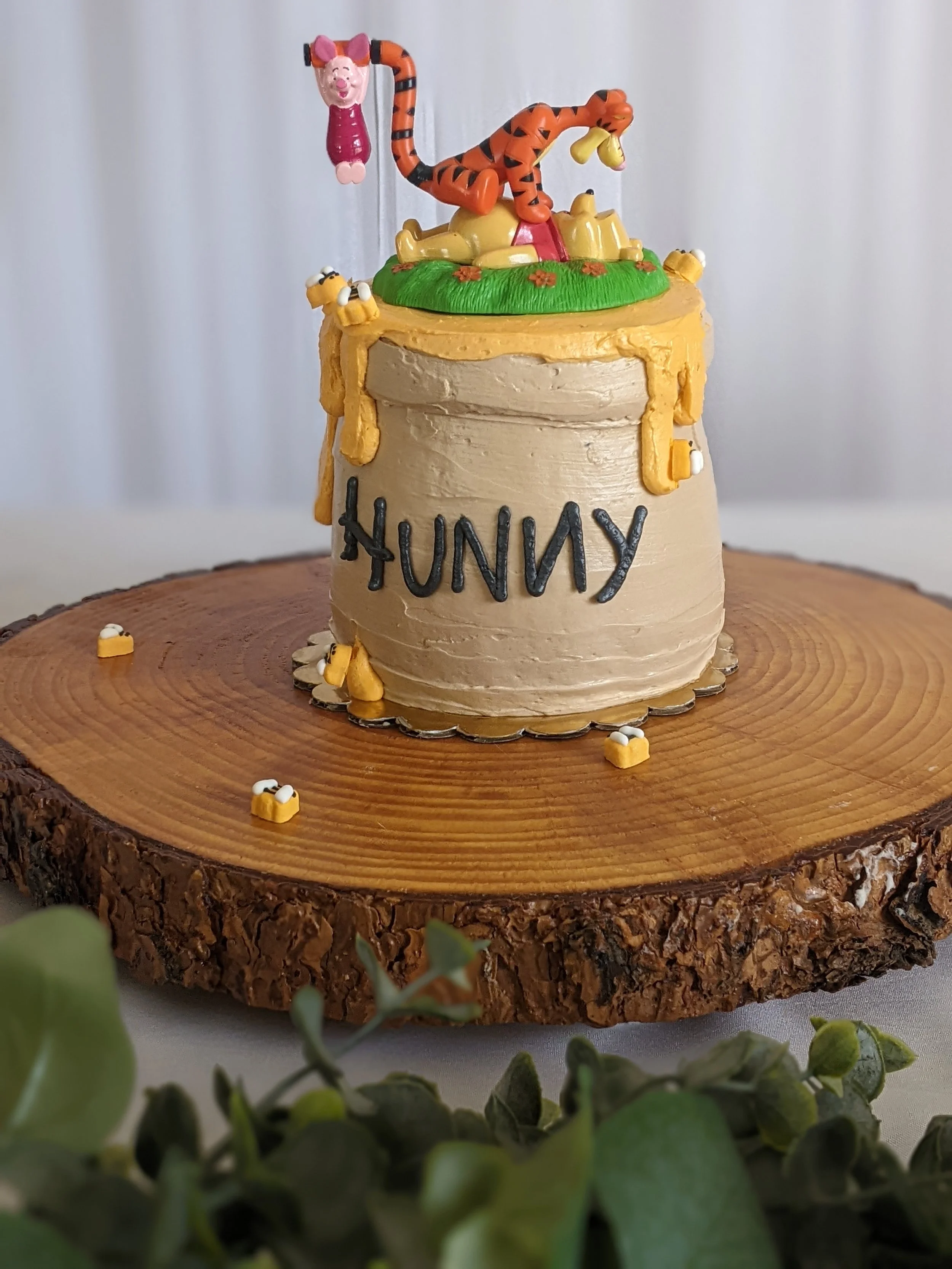 A decorated birthday cake on a wooden slab with a Winnie the Pooh theme, featuring a Tigger topper and honey pot decorations, with the name 'HUNNY' written on the side.