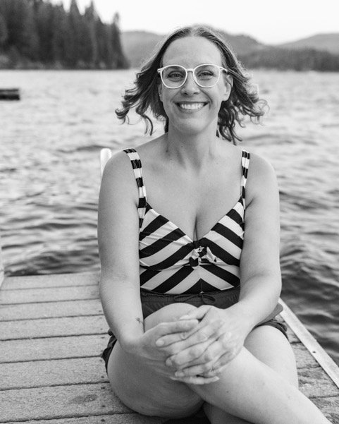 Woman sitting on a dock by a lake, wearing glasses and a striped sleeveless top, smiling at the camera.