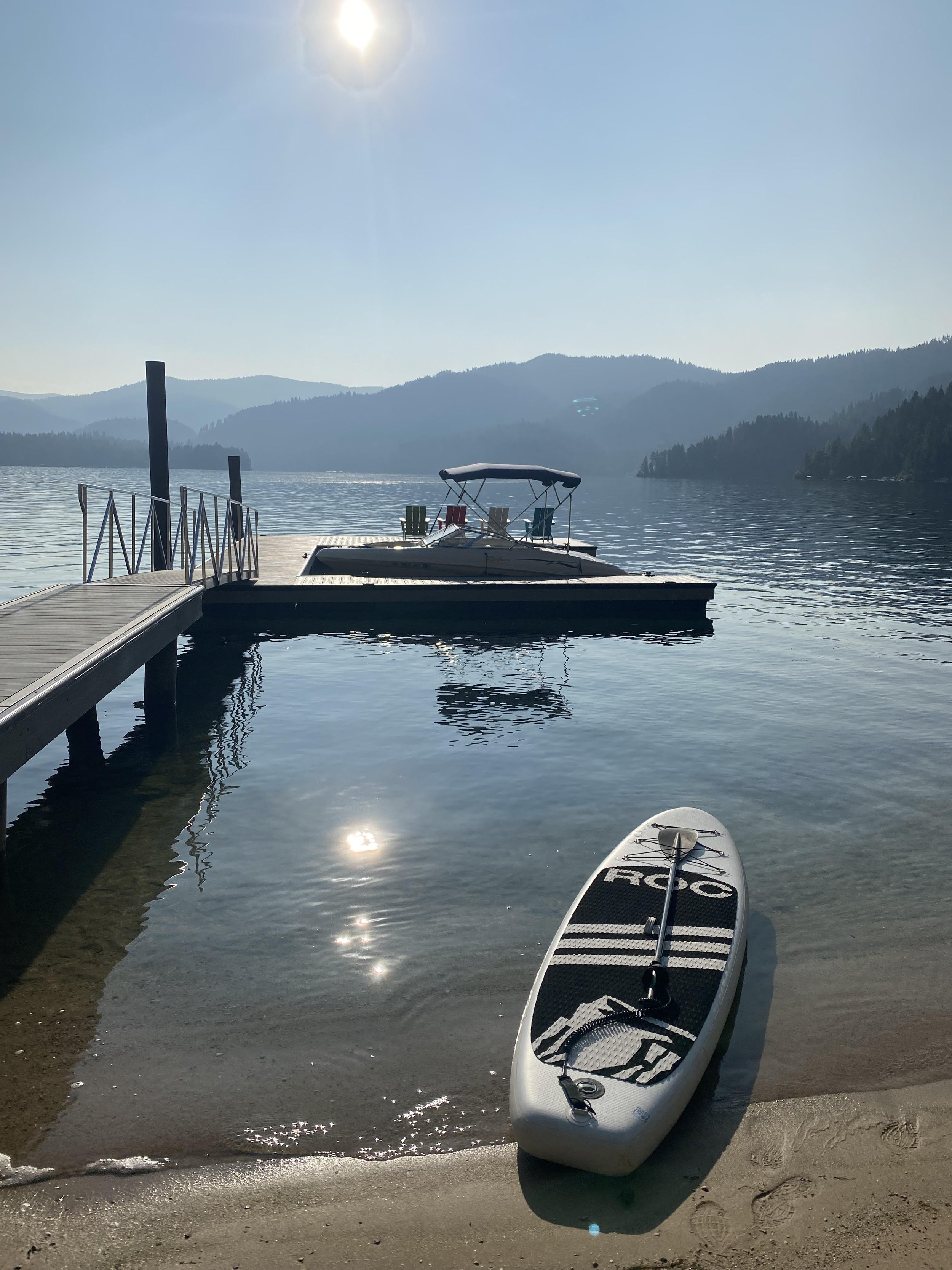 A paddleboard on the sandy shore of a lake with a dock, boats on the water, and mountains in the background under a sunny sky.
