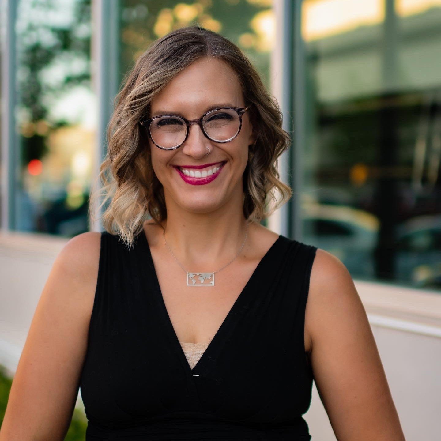 A woman with shoulder-length curly hair, glasses, and a big smile, wearing a black sleeveless top and a silver necklace, standing outdoors in front of a building with large windows.