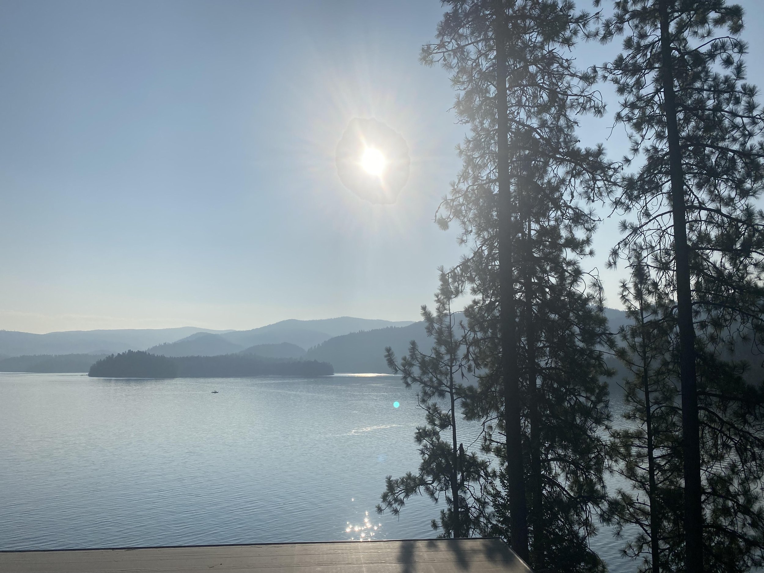 Serene lake with small boat, surrounded by mountainous landscape, tall pine trees on right, bright sun in clear blue sky.