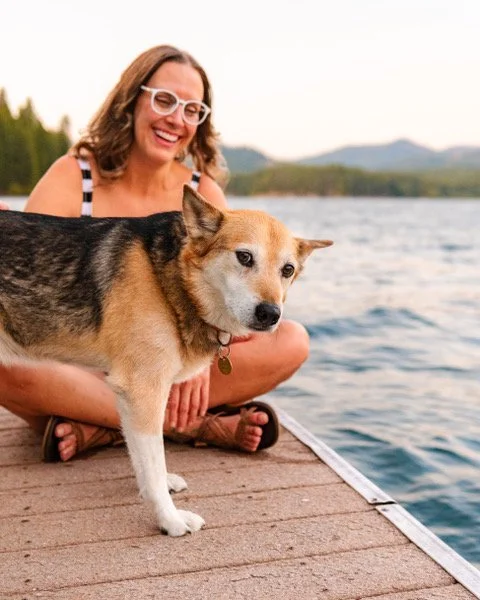 A woman with glasses smiling and sitting by a lake while holding a dog on a dock.