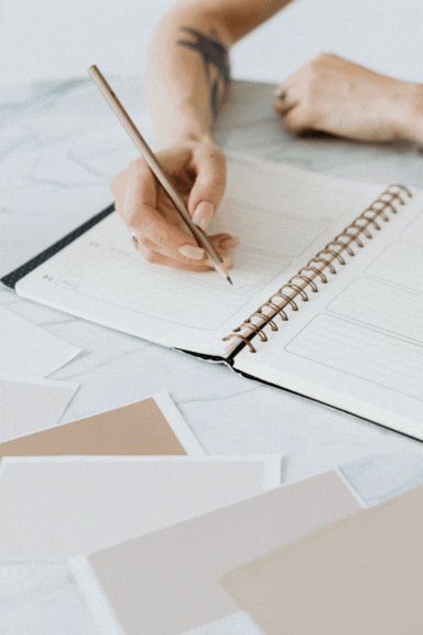 A person writing in a spiral notebook with a pencil, surrounded by color swatches on a marble surface.