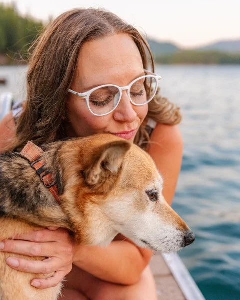 A woman wearing glasses tenderly hugging her dog near a body of water with trees and mountains in the background.