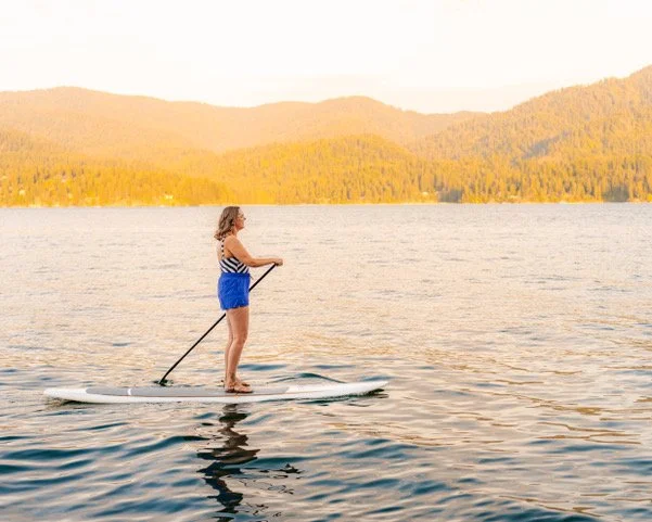 A woman paddleboarding on a lake with mountains in the background during sunset.