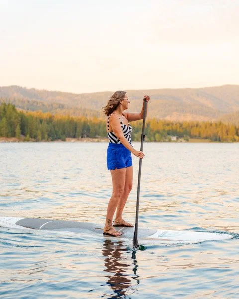 Woman paddleboarding on a lake with a mountainous backdrop and forested shoreline.