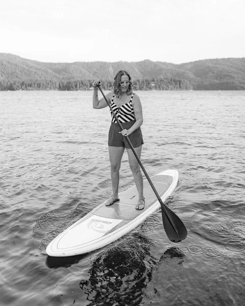 Woman standing on paddleboard with paddle on a lake, wearing sunglasses, a zigzag bikini top, and shorts, with forested hills in the background.