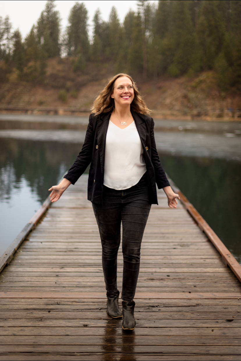 A woman walking on a wooden dock over a body of water, smiling with her arms slightly outstretched, surrounded by forested hills.