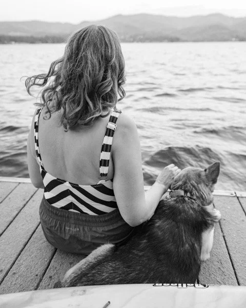 A woman with wavy hair sitting on a dock by a lake, holding a dog while looking at the water in black and white.