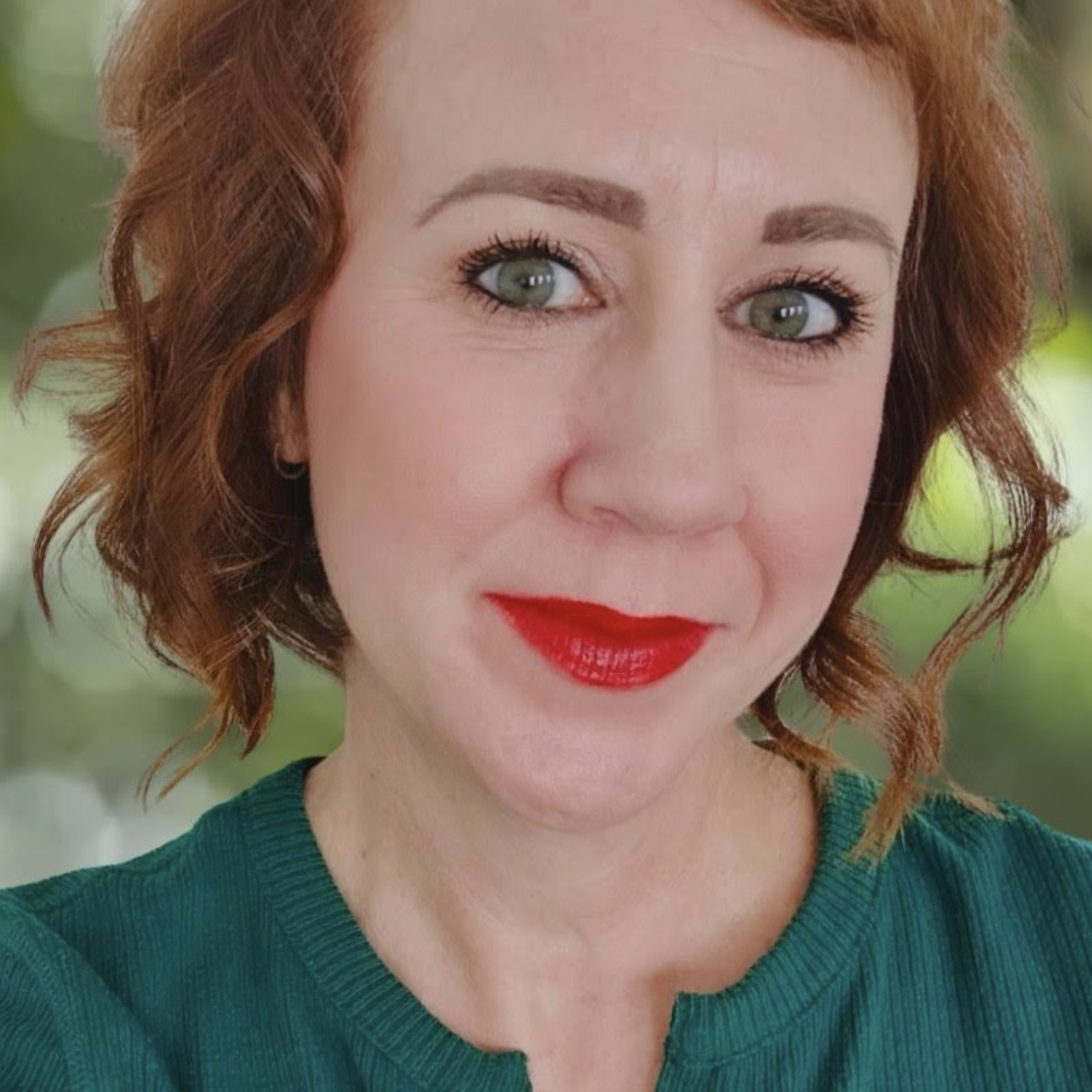 Close-up of a woman with red hair, wearing red lipstick and a green top, with a blurred green background.