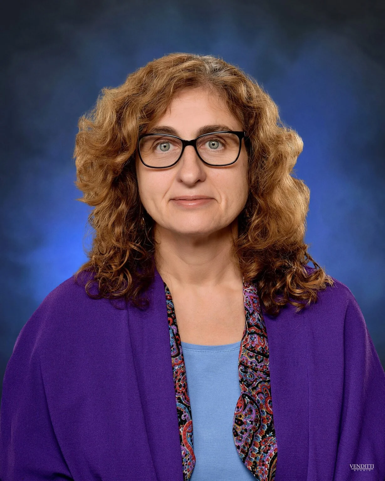 A woman with curly red hair wearing glasses, a purple blazer, and a patterned scarf, posing against a blue background.