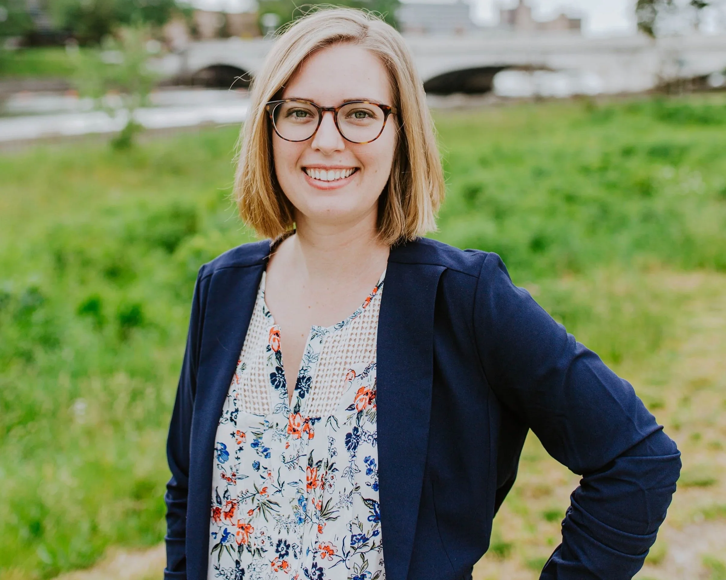 A woman with shoulder-length blonde hair, glasses, and a friendly smile standing outdoors in a green park, wearing a navy blazer over a floral patterned blouse.