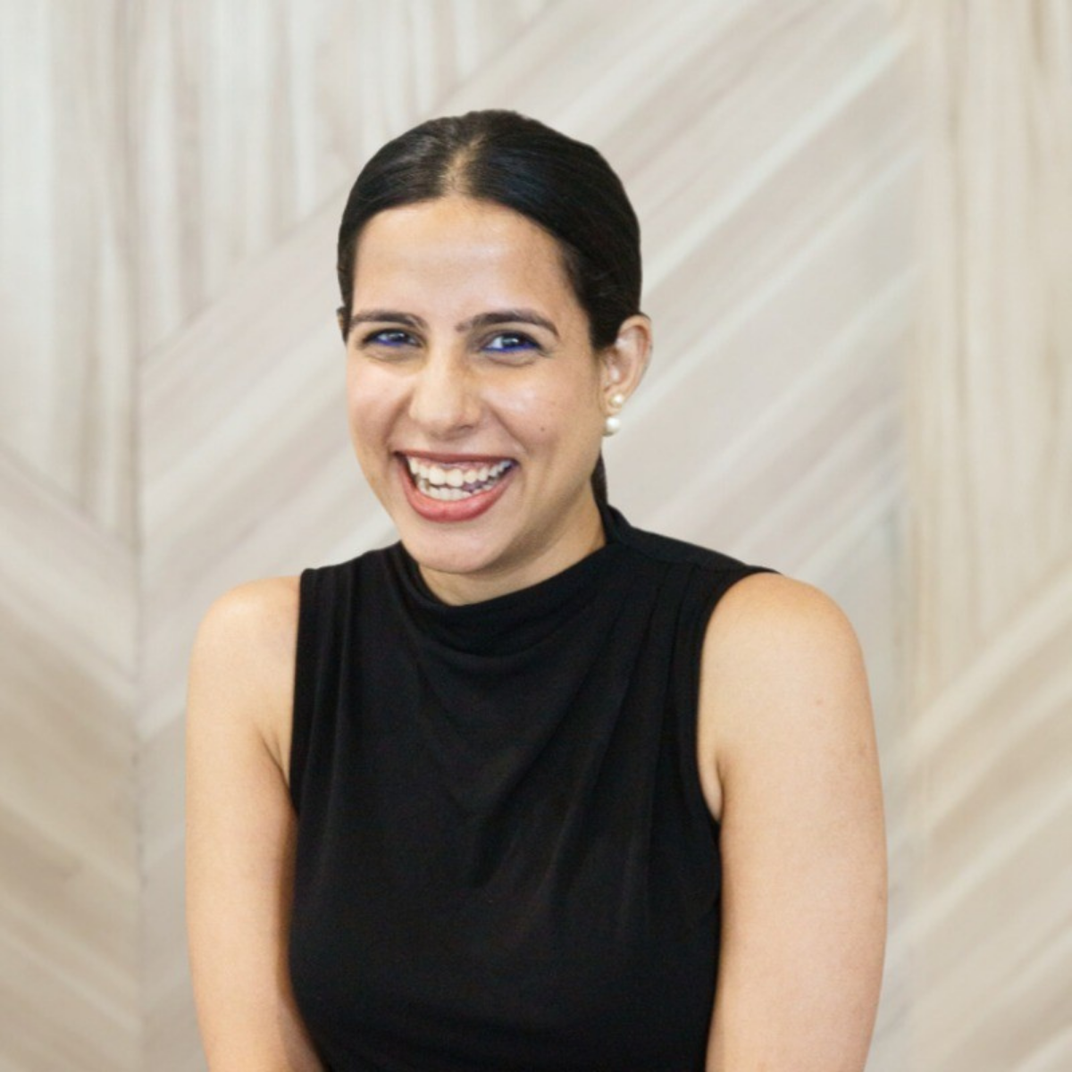 A woman with dark hair in a bun, wearing pearl earrings and a sleeveless black top, smiling in front of a light wood-paneled wall.