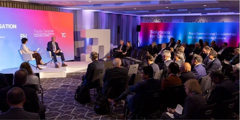 A professional conference with a stage featuring a large screen, two speakers seated on stage, and an audience of business professionals seated and attentively listening in a dimly lit conference room with purple and pink lighting.