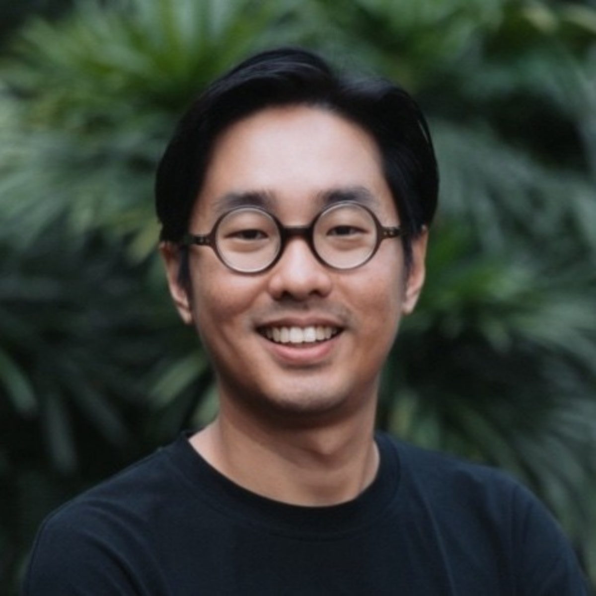 A smiling young Asian man with black hair, round glasses, wearing a black T-shirt, standing outdoors with green foliage in the background.