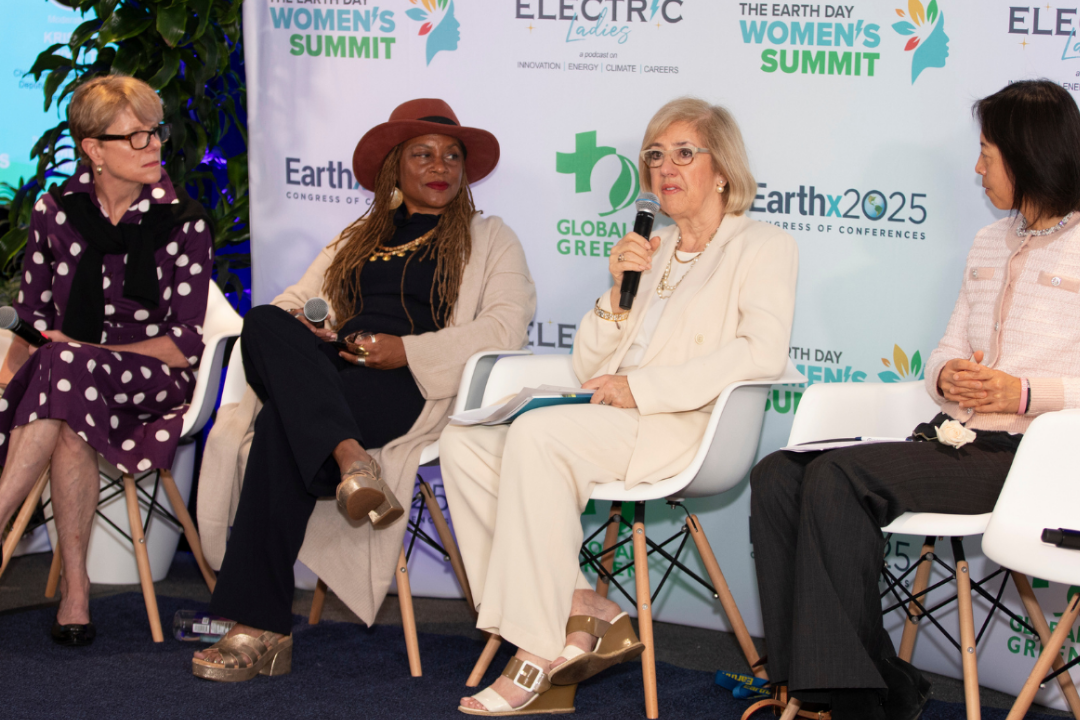 Four women seated on stage at a panel discussion during the Earth Day Women's Summit, with a backdrop displaying Earth Day and environmental organization logos, and one woman speaking into a microphone.