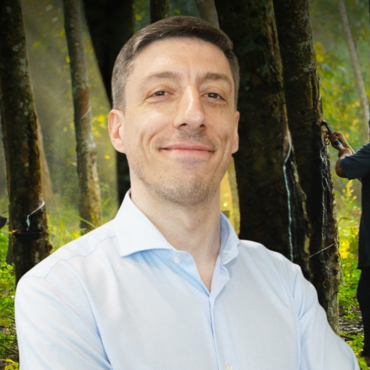 A man in a light blue shirt smiling in a forest with blurred trees and a person in the background pouring water from a bottle.