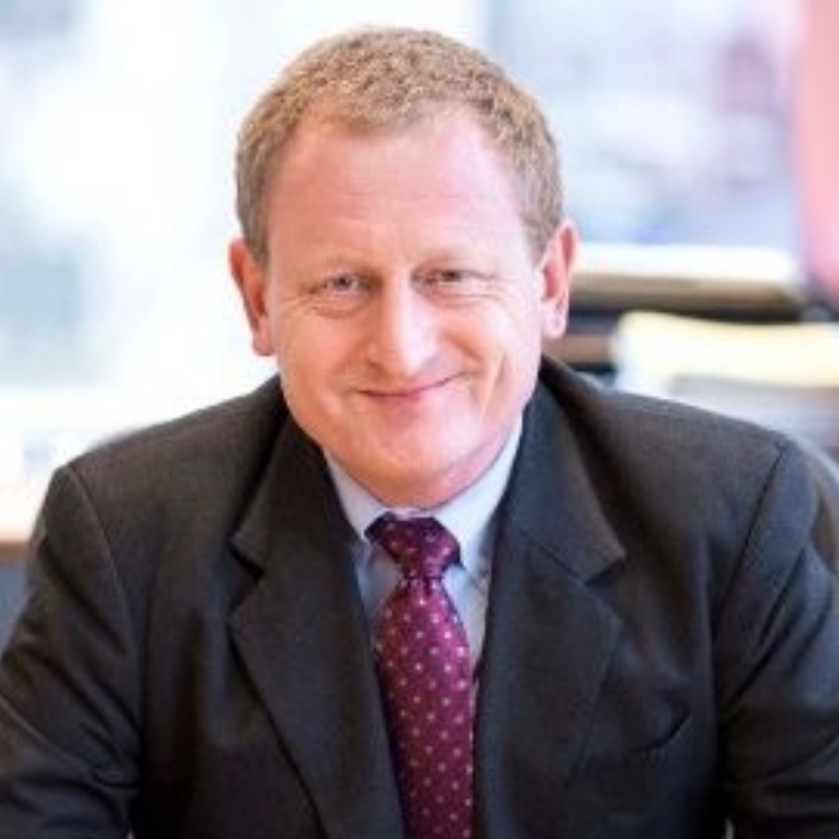 A middle-aged man with red hair in a suit and purple tie, smiling in an office setting.