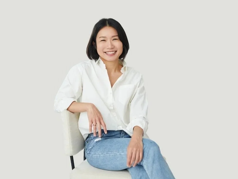 A young woman with short black hair smiling, wearing a white shirt and blue jeans, sitting on a chair with a plain, light-colored background.