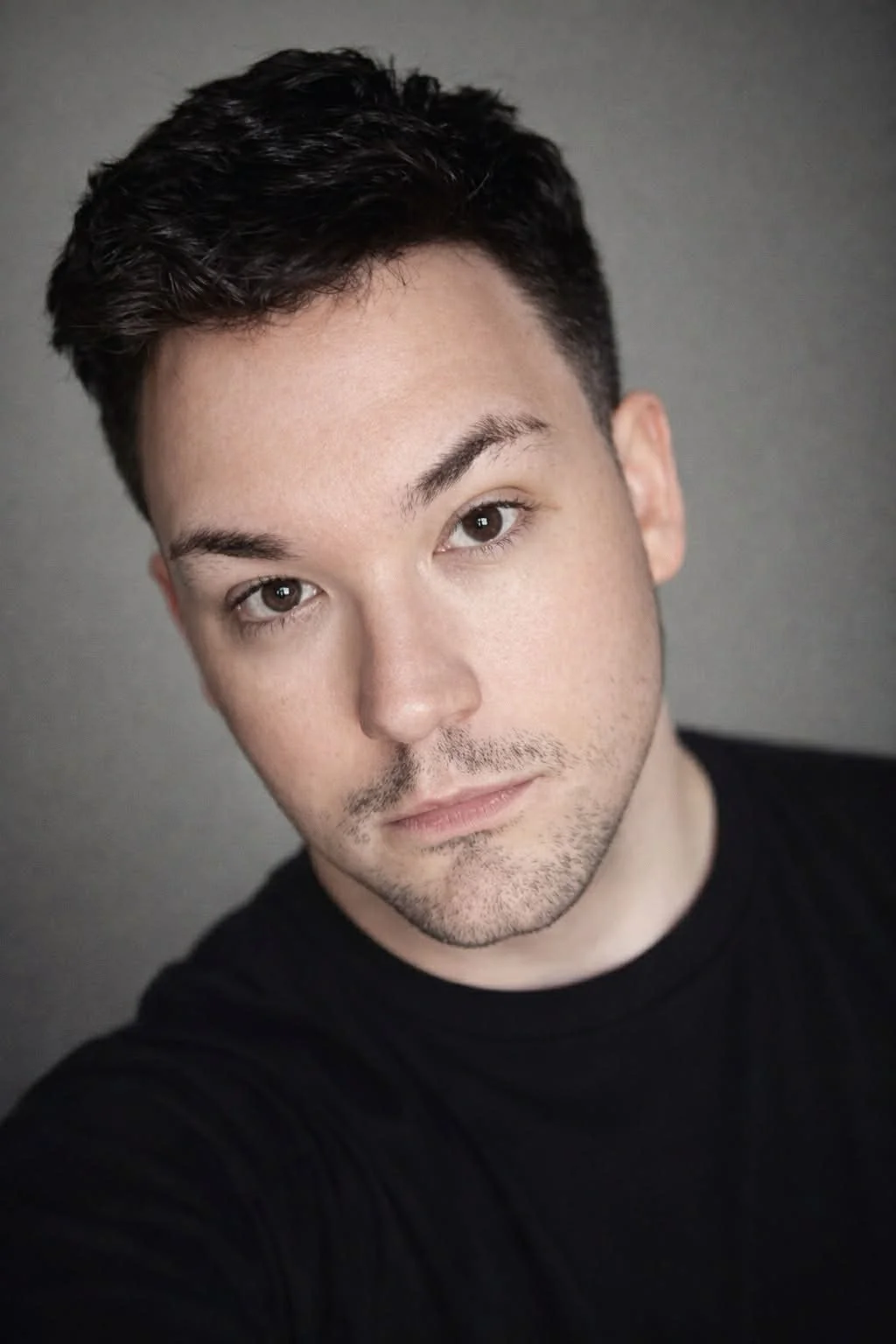 Close-up portrait of a young man with short dark hair, brown eyes, and a light beard, wearing a black shirt, against a gray background.