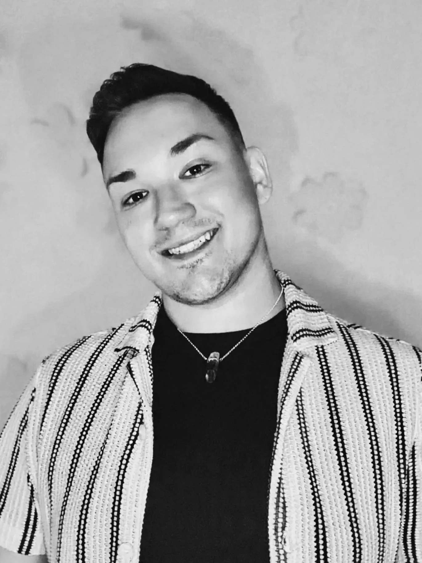Black and white photo of a young man with short, dark hair, smiling, wearing a striped shirt over a dark t-shirt with a necklace, against a plain wall.