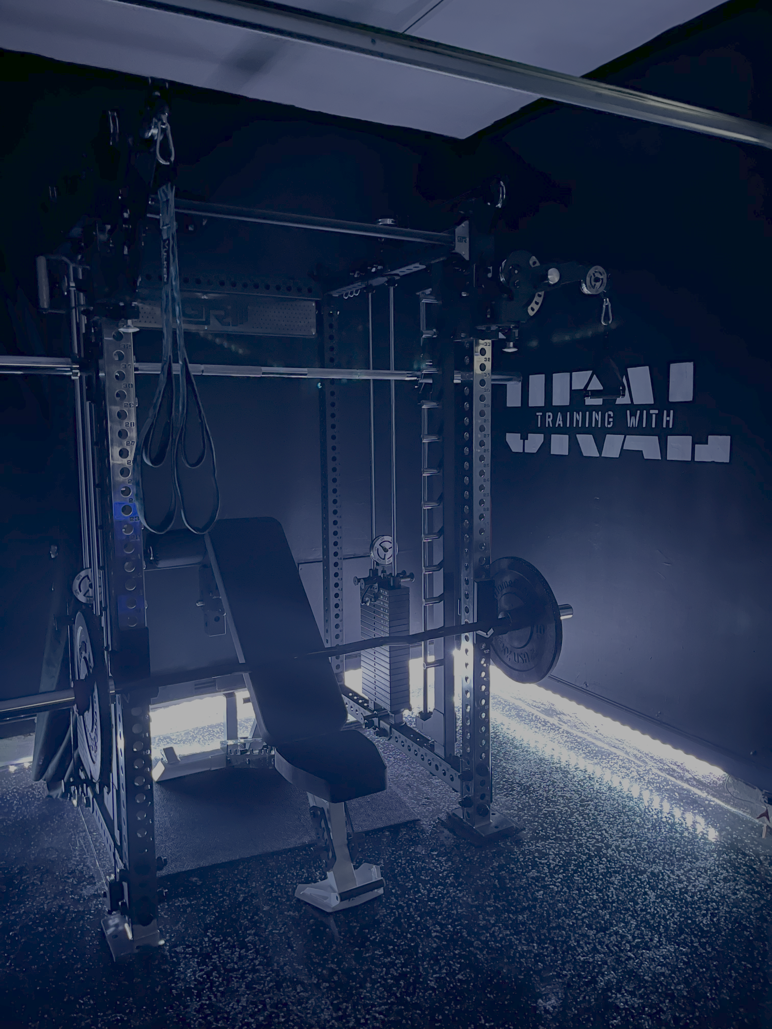 A home gym with black walls, a large metal squat rack, a weight bench, and various weights and gym equipment illuminated by blue LED lights.