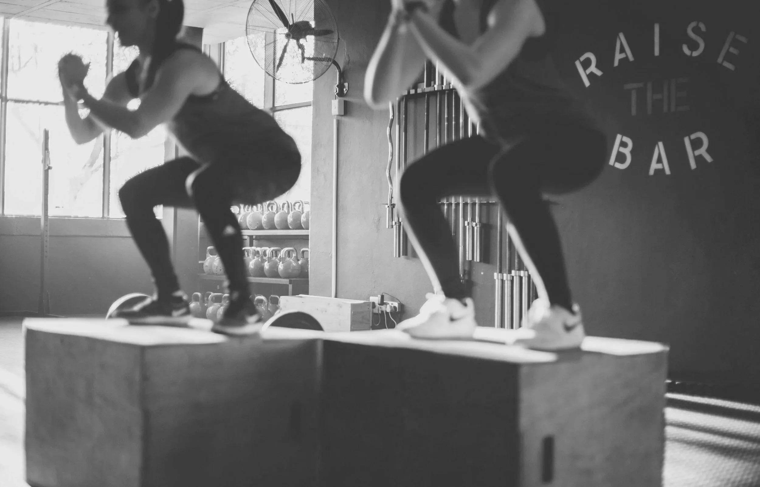 Two people working out with kettlebells on an elevated platform in a gym, with kettlebells on shelves in the background.