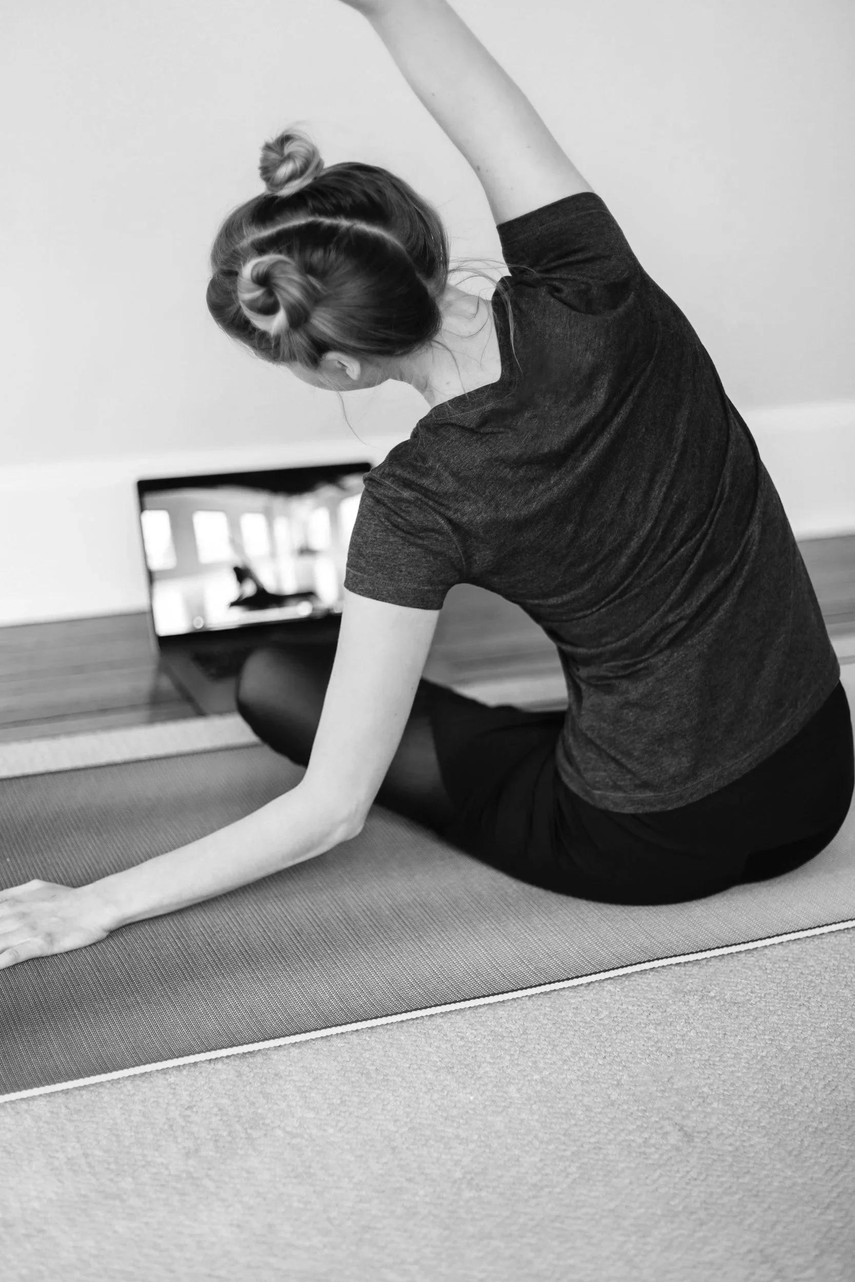 A woman in workout clothes demonstrates stretches on a yoga mat while watching a fitness class on a laptop.