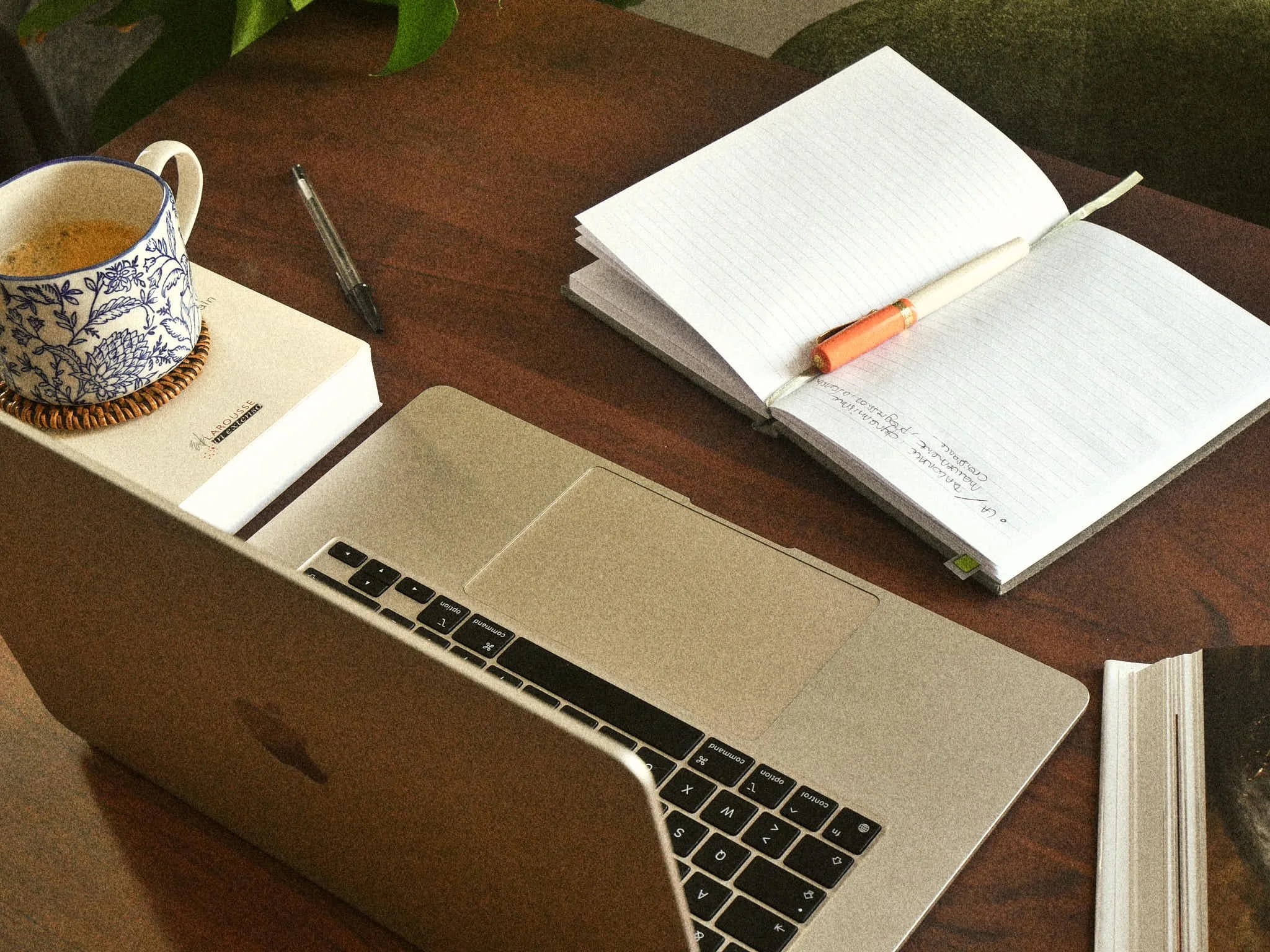 Un bureau avec un ordinateur portable, un carnet ouvert avec un stylo orange, une tasse avec une boisson chaude, un livre, un marque-page, un stylo noir, et un paquet de biscuits.